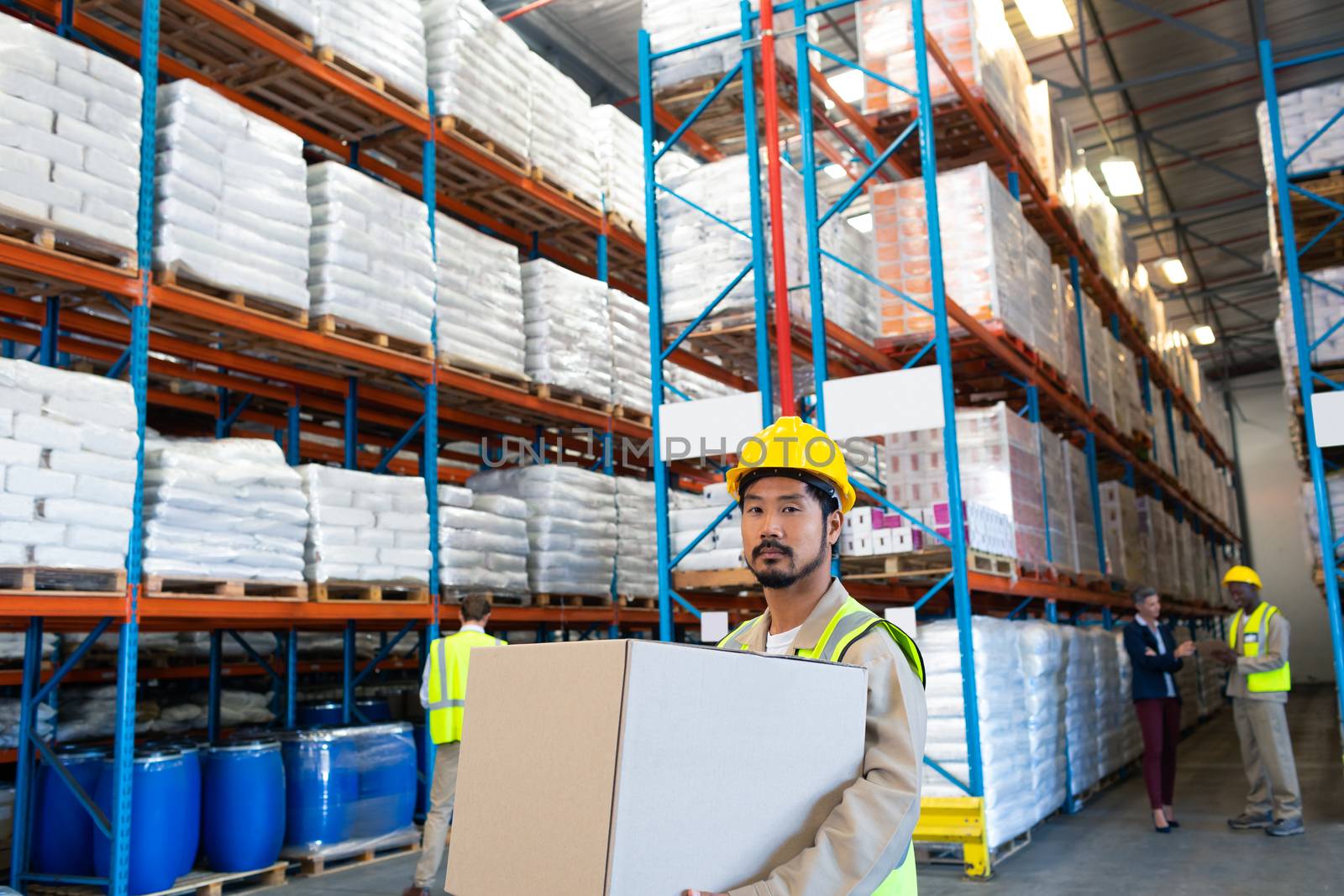 Front view of handsome mature Asian male worker carrying cardboard box and looking at camera in warehouse. This is a freight transportation and distribution warehouse. Industrial and industrial workers concept
