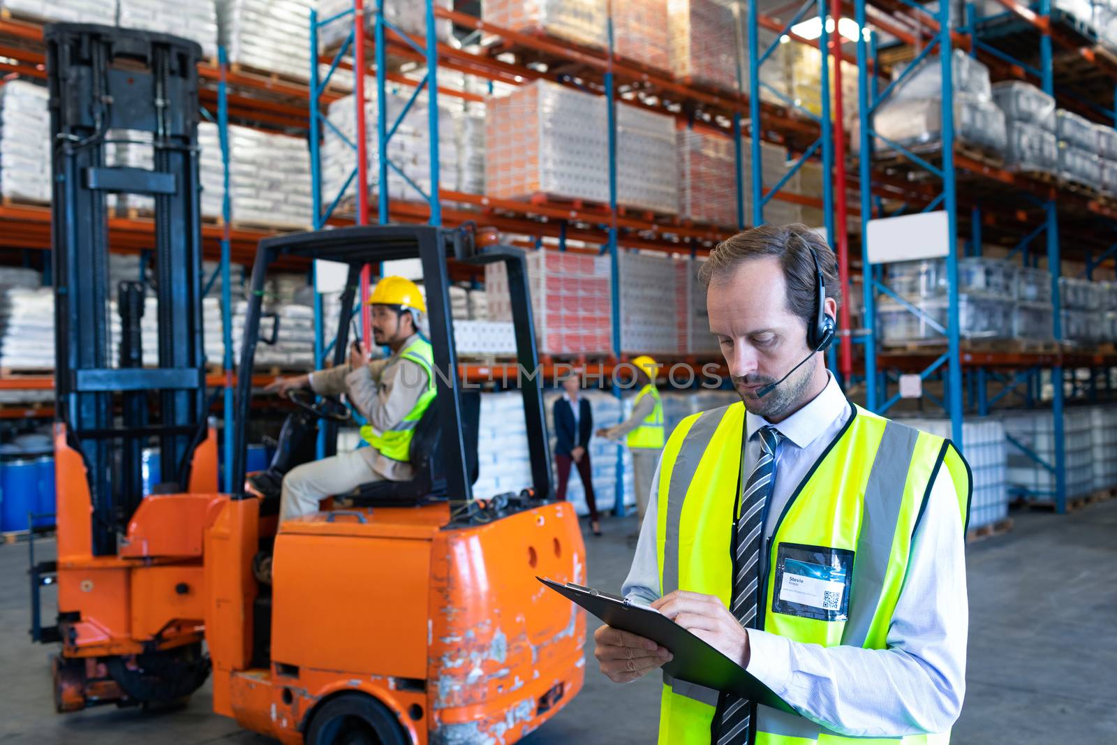Front view of handsome Caucasian male supervisor with headset writing on clipboard in warehouse. This is a freight transportation and distribution warehouse. Industrial and industrial workers concept