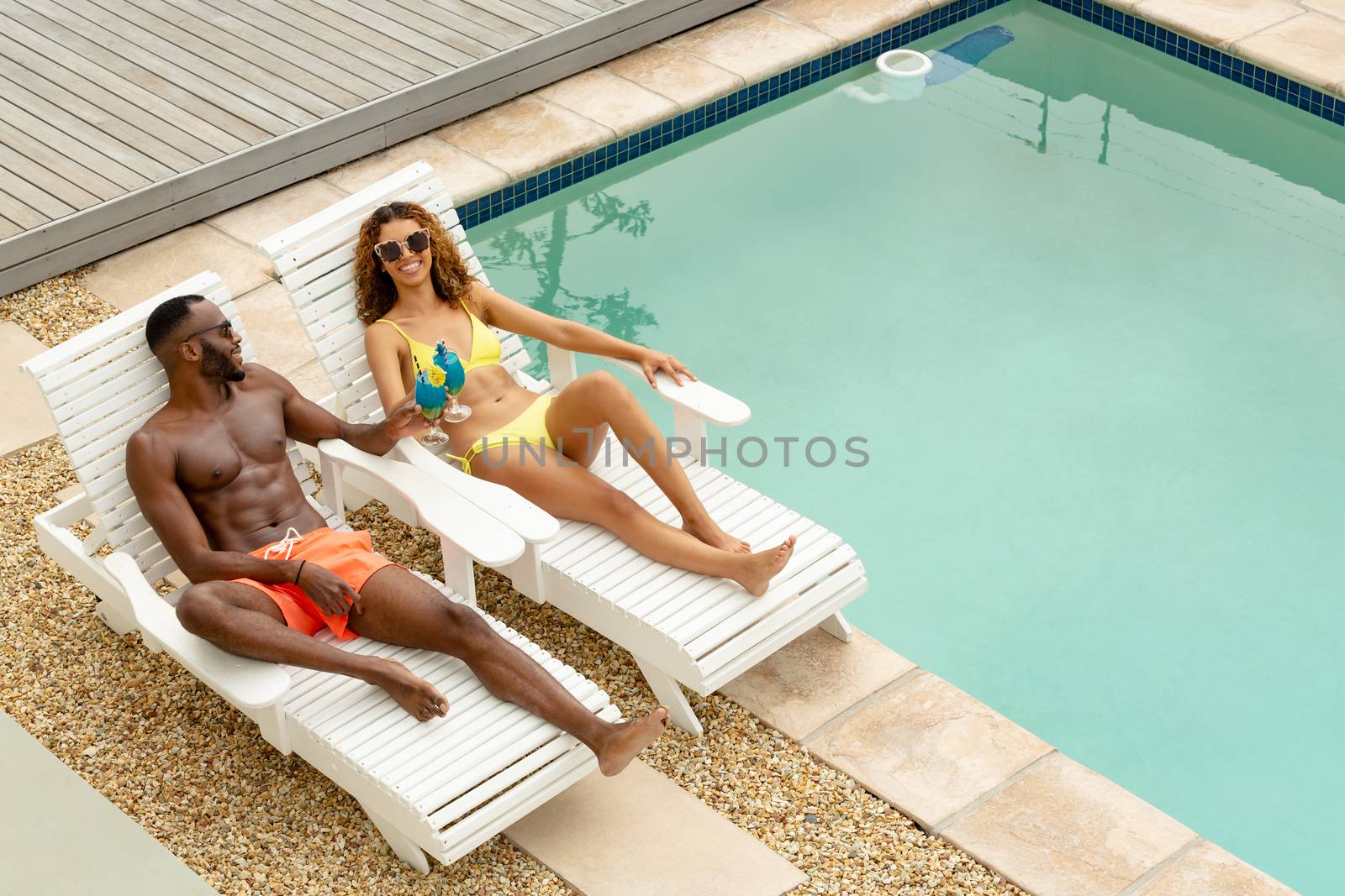 High angle view of cute diverse couple toasting glasses of cocktail while relaxing on a sun lounger near swimming pool at the backyard. Summer fun at home by the swimming pool