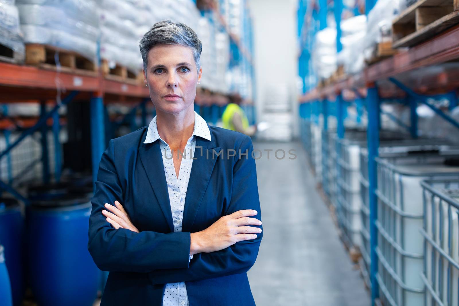 Portrait close-up of beautiful mature Caucasian female manager standing with arm crossed and looking at camera in warehouse. African-american colleague standing in the background. This is a freight transportation and distribution warehouse. Industrial and industrial workers concept