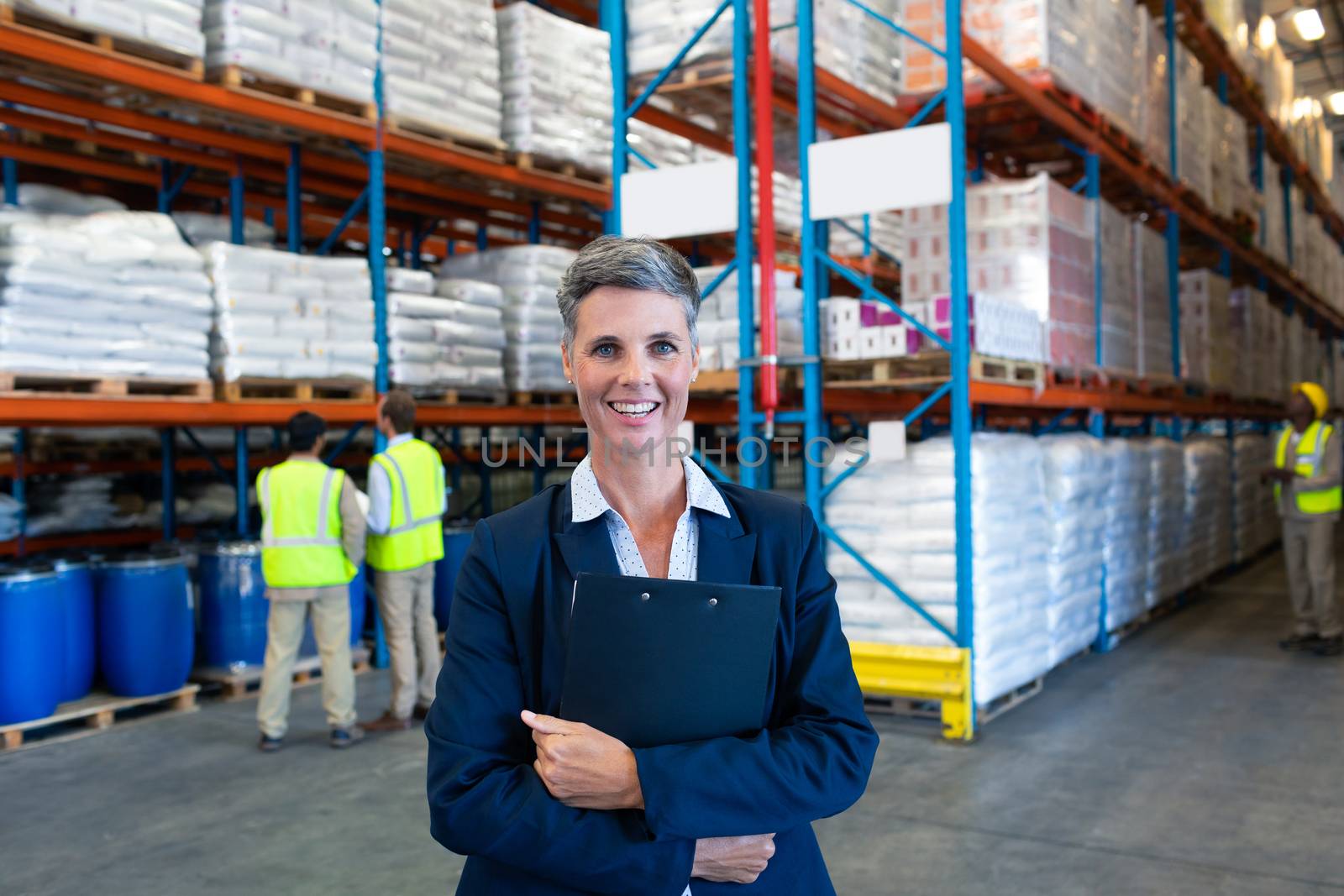 Front view of mature Caucasian female manager holding clipboard and looking at camera in warehouse. This is a freight transportation and distribution warehouse. Industrial and industrial workers concept