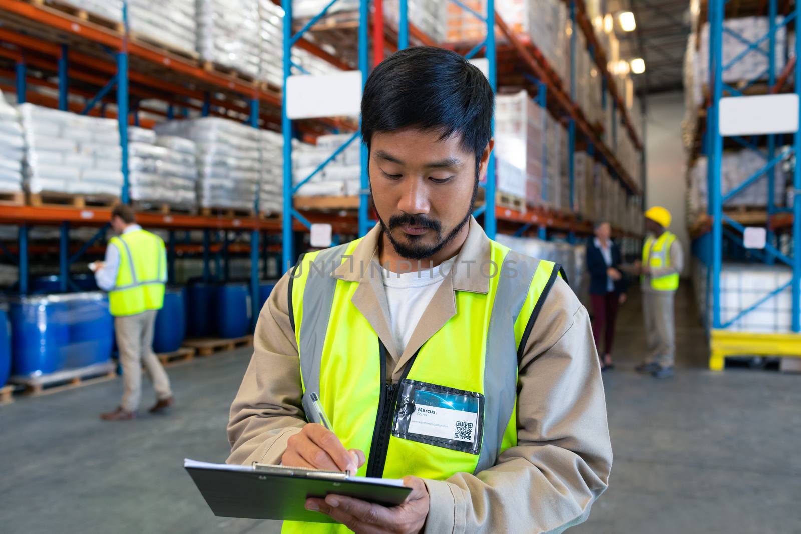 Close-up of handsome mature Asian male worker writing on clipboard in warehouse. Diverse colleagues working in the background. This is a freight transportation and distribution warehouse. Industrial and industrial workers concept
