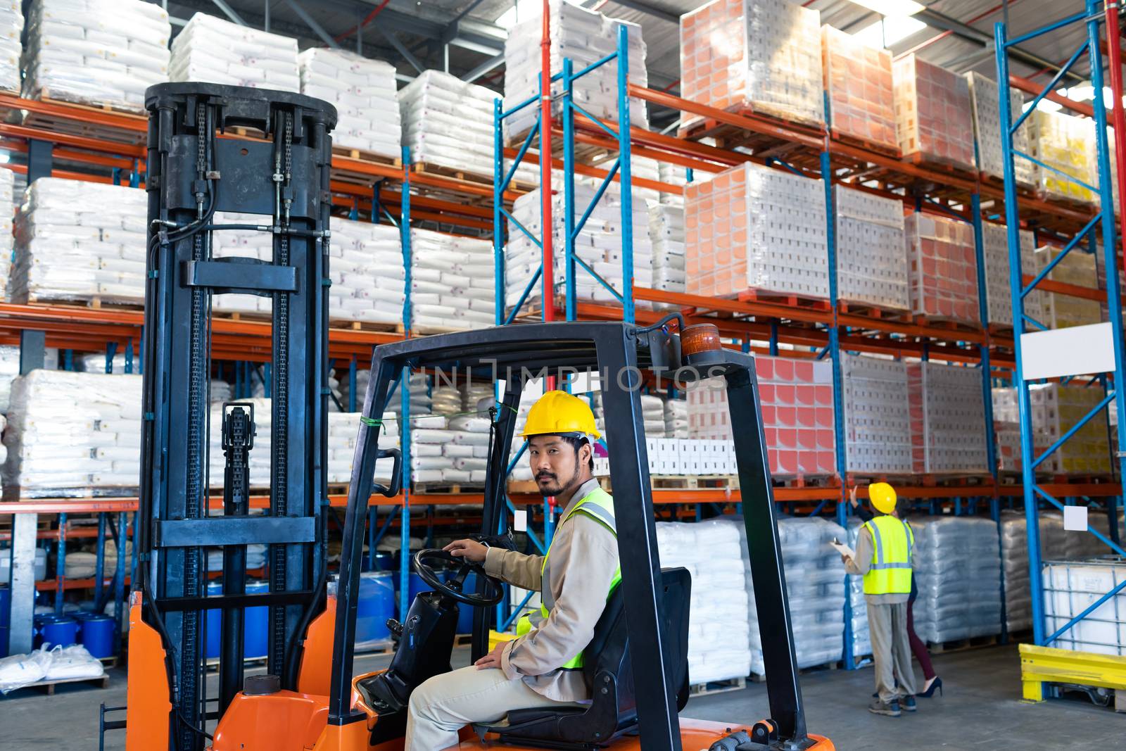 Side view of handsome mature Asian male worker sitting in forklift and looking at camera in warehouse. Diverse colleagues working in the background. This is a freight transportation and distribution warehouse. Industrial and industrial workers concept
