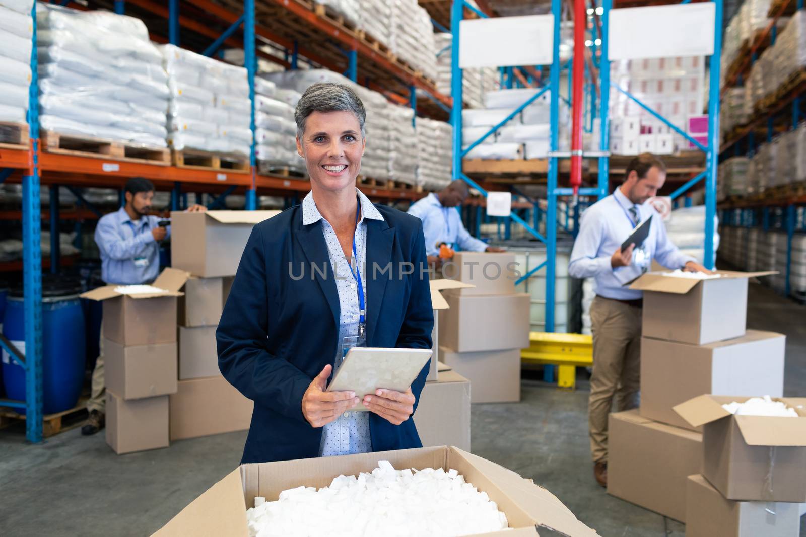 Front view of happy mature Caucasian female manager with digital tablet looking at camera in warehouse. Diverse mature warehouse worker unpacking cardboard boxes. This is a freight transportation and distribution warehouse. Industrial and industrial workers concept
