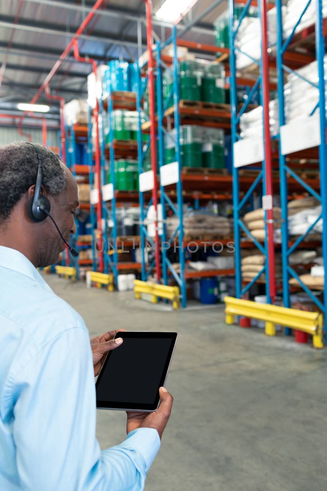 Rear view of mature African-american male supervisor with headset using digital tablet in warehouse. This is a freight transportation and distribution warehouse. Industrial and industrial workers concept