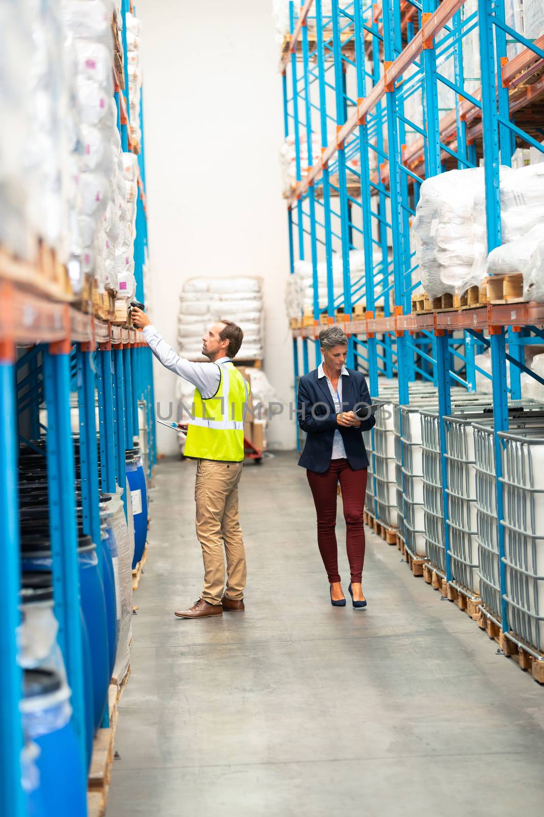 Side view of handsome mature Caucasian male worker scanning package with barcode scanner in modern warehouse. Caucasian woman is walking with clipboard in aisle behind male worker. This is a freight transportation and distribution warehouse. Industrial and industrial workers concept