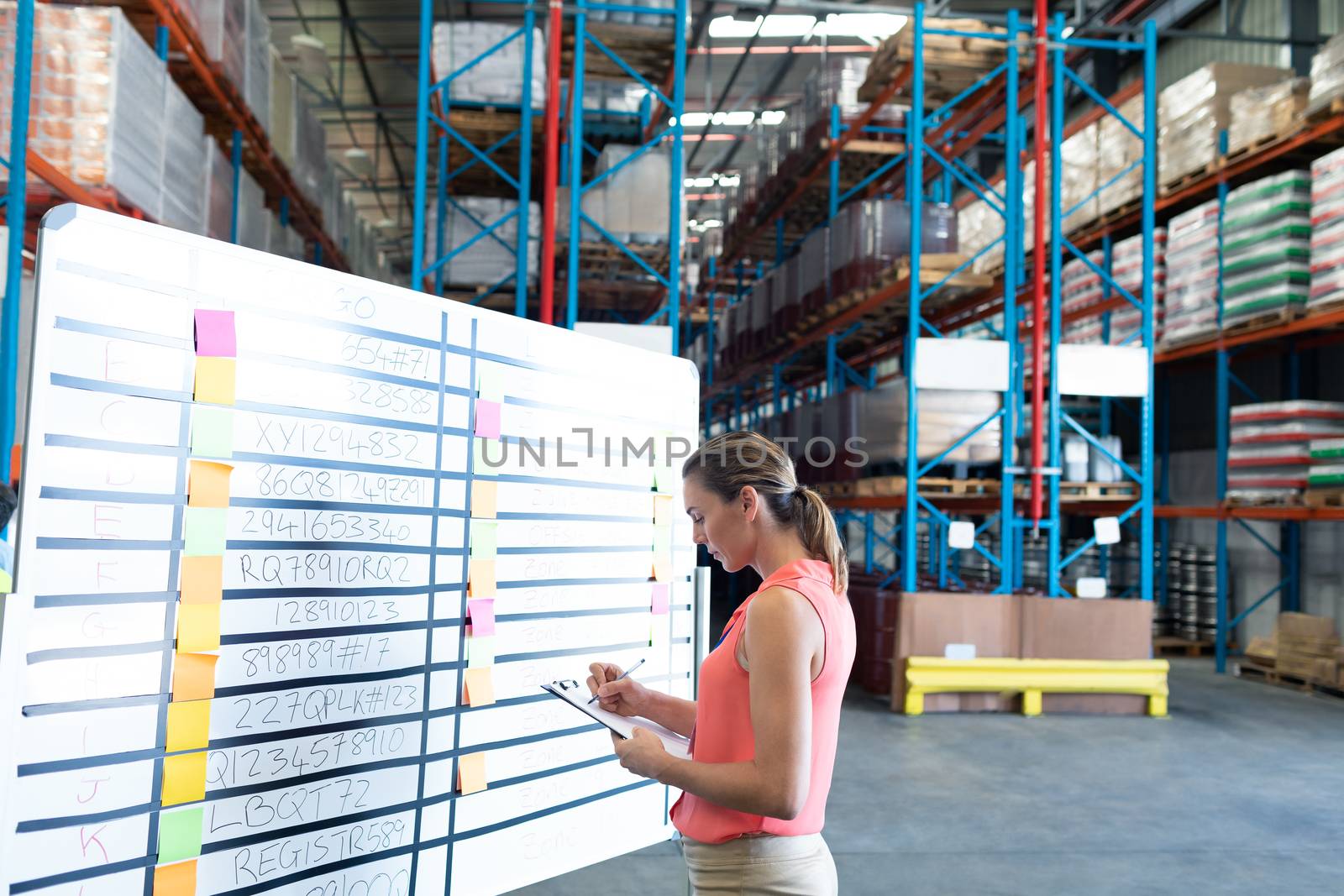 Side view of pretty young Caucasian female staff writing on clipboard near whiteboard in warehouse. This is a freight transportation and distribution warehouse. Industrial and industrial workers concept