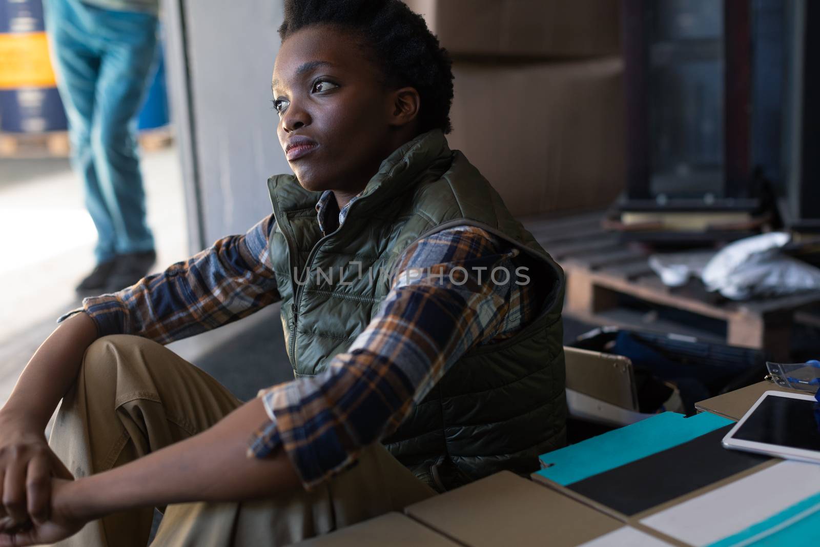 Side view of African-american thoughtful female worker sitting in warehouse. This is a freight transportation and distribution warehouse. Industrial and industrial workers concept