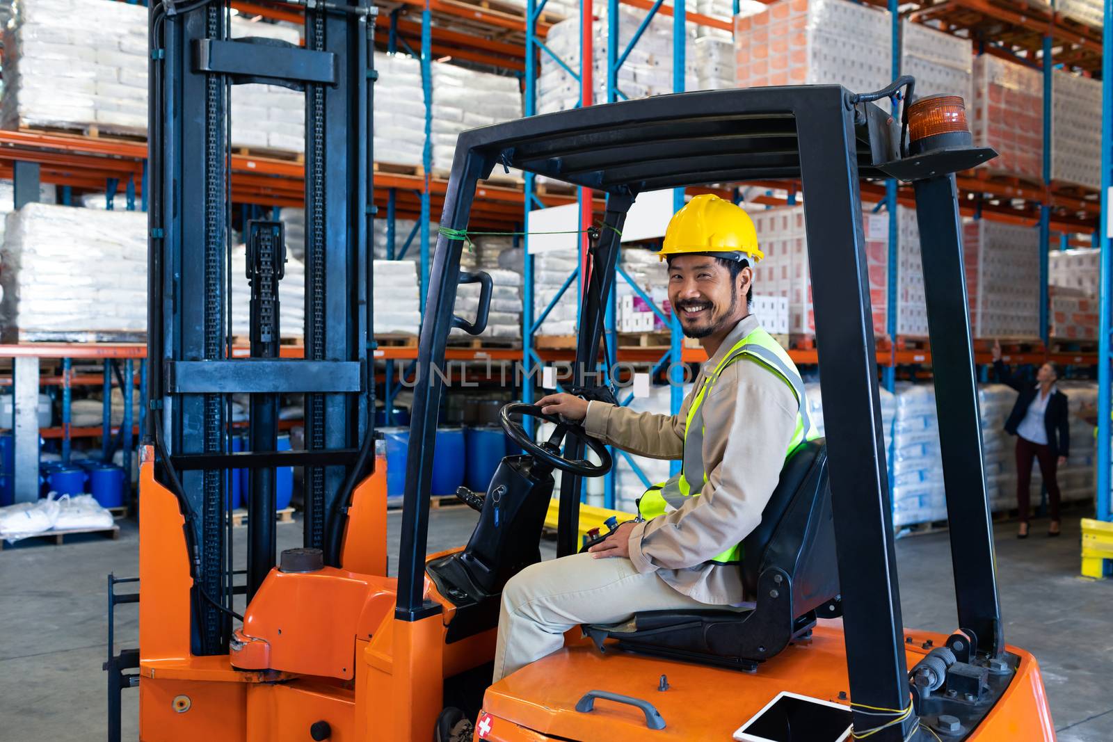 Front view of happy mature Asian male worker sitting in forklift and looking at camera in warehouse. This is a freight transportation and distribution warehouse. Industrial and industrial workers concept