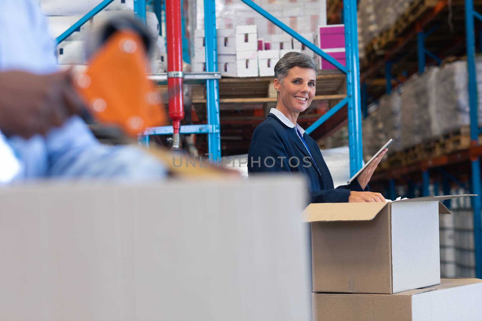 Side view of happy mature Caucasian female manager looking at camera while working on digital tablet in warehouse. African-american male worker works on the foreground. This is a freight transportation and distribution warehouse. Industrial and industrial workers concept