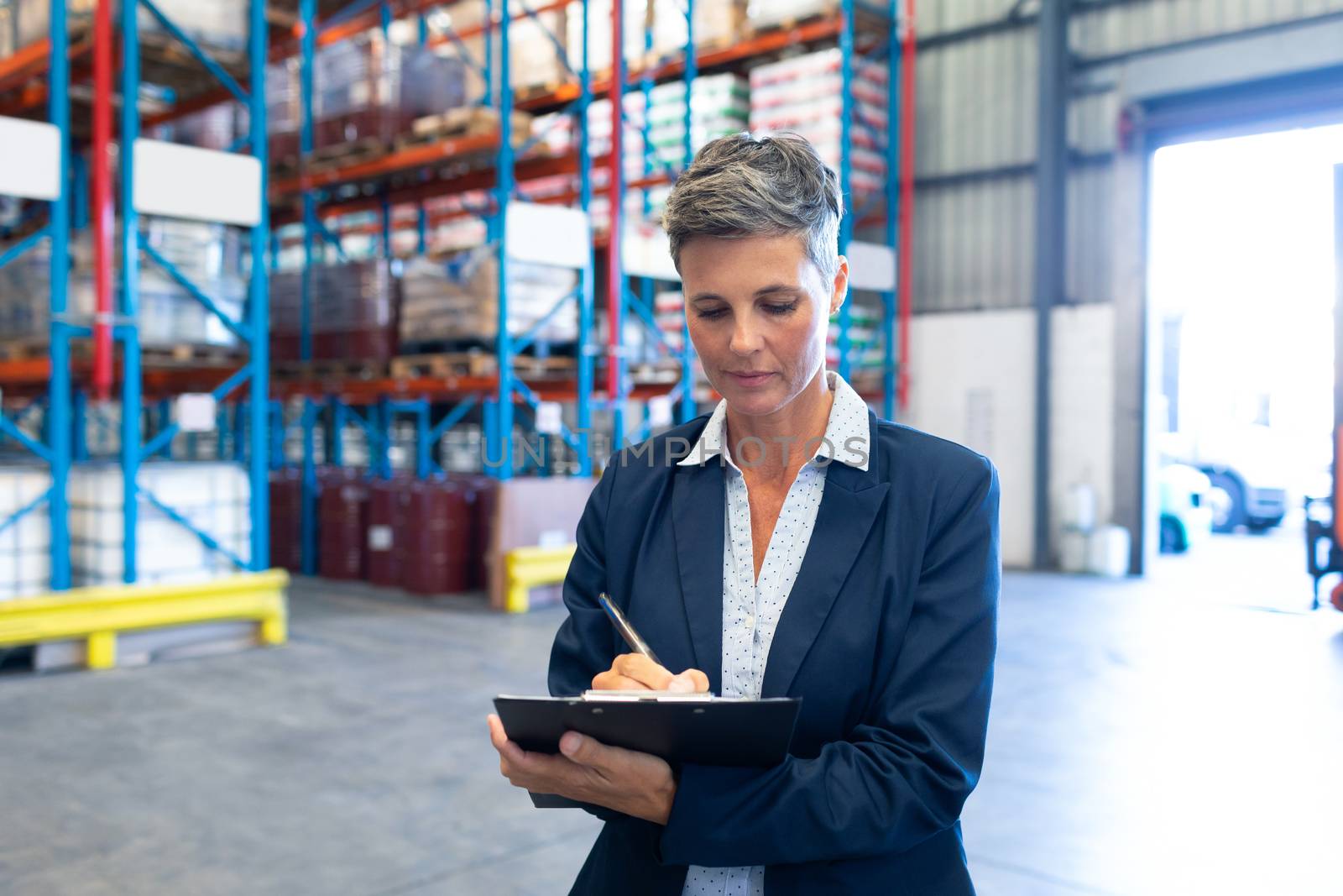 Front view of beautiful mature Caucasian female manager writing on clipboard in warehouse. This is a freight transportation and distribution warehouse. Industrial and industrial workers concept