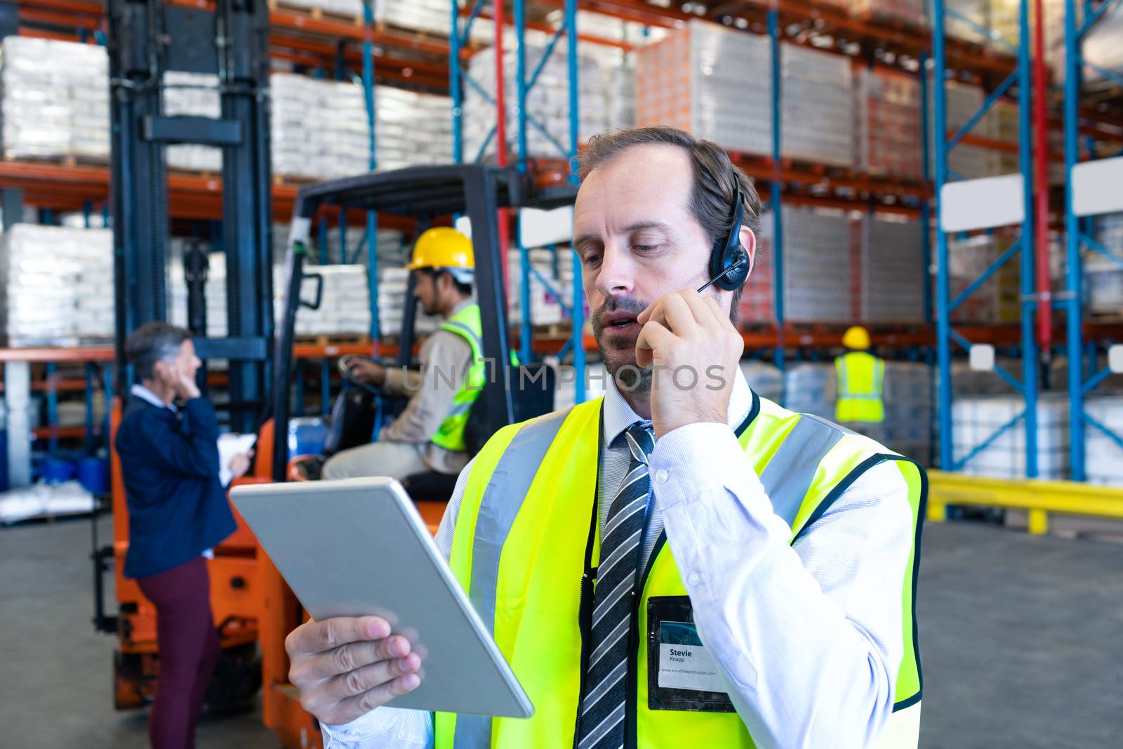 Close-up of handsome Caucasian male supervisor using digital tablet while talking on headset in warehouse. Diverse colleagues communicating in the background. This is a freight transportation and distribution warehouse. Industrial and industrial workers concept