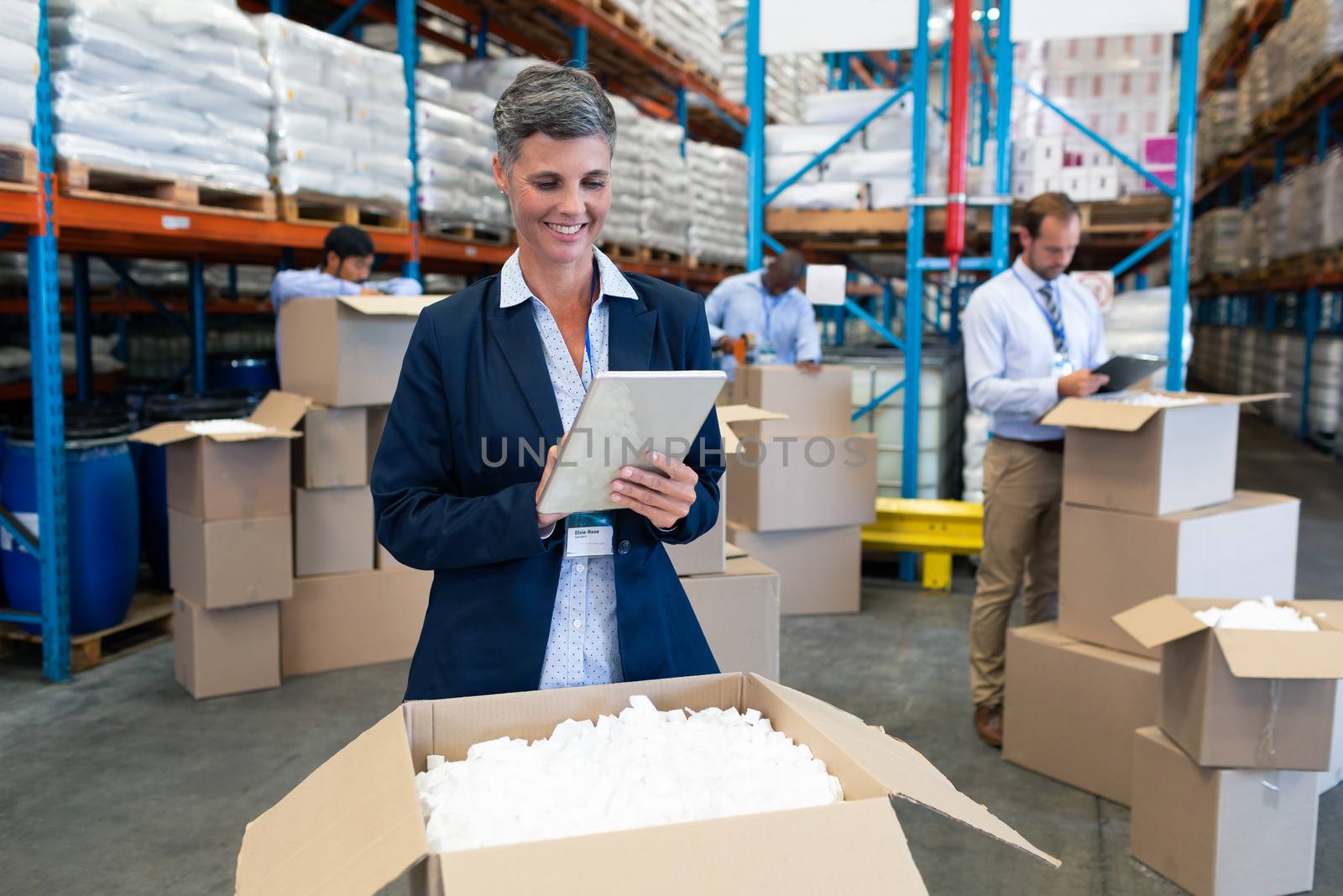 Front view of happy Caucasian female manager working on digital tablet in warehouse. Diverse warehouse workers unpacking behind her. This is a freight transportation and distribution warehouse. Industrial and industrial workers concept