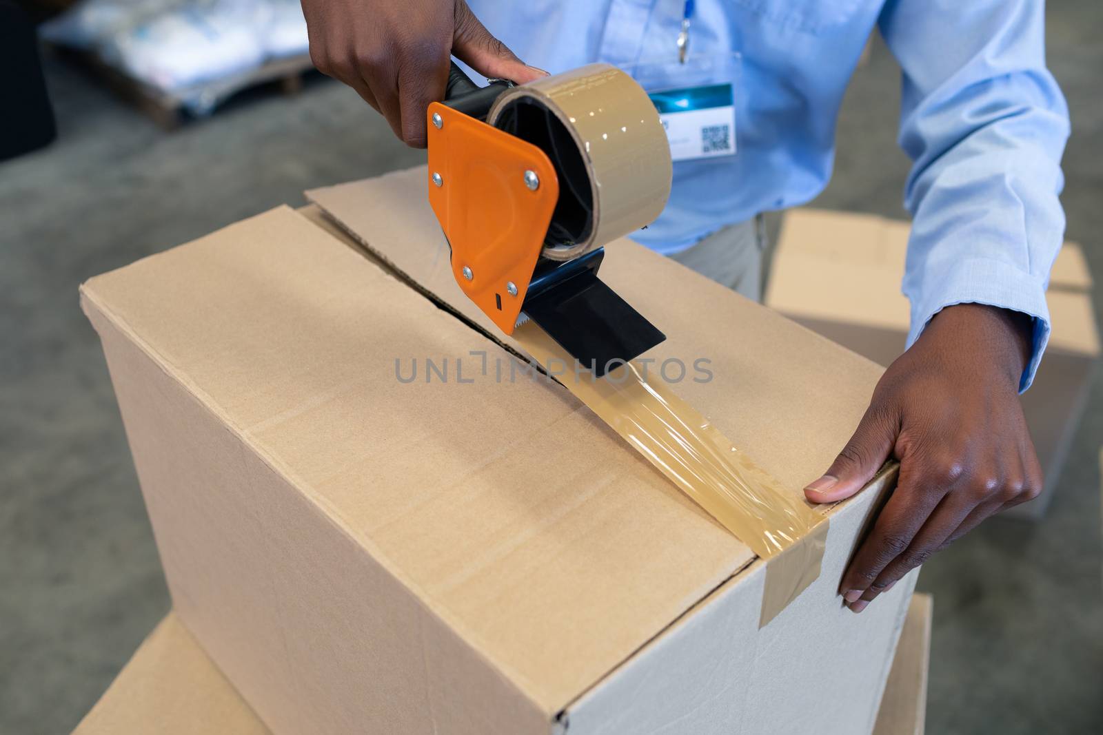 Close-up of mature Caucasian male staff packing cardboard box with tape gun dispenser in warehouse. This is a freight transportation and distribution warehouse. Industrial and industrial workers concept