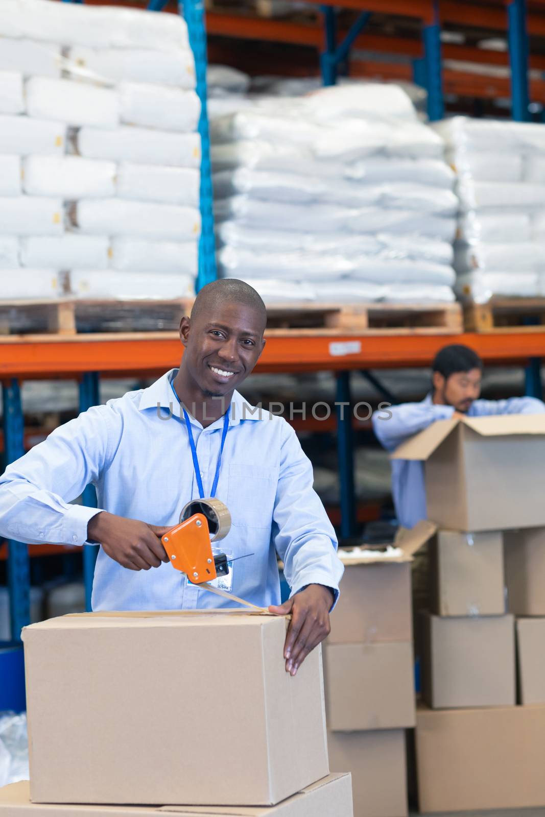 Portrait close-up of happy mature African-american male staff packing cardboard box with tape gun dispenser in warehouse. Asian male worker is unpacking cardboard box in the background. This is a freight transportation and distribution warehouse. Industrial and industrial workers concept