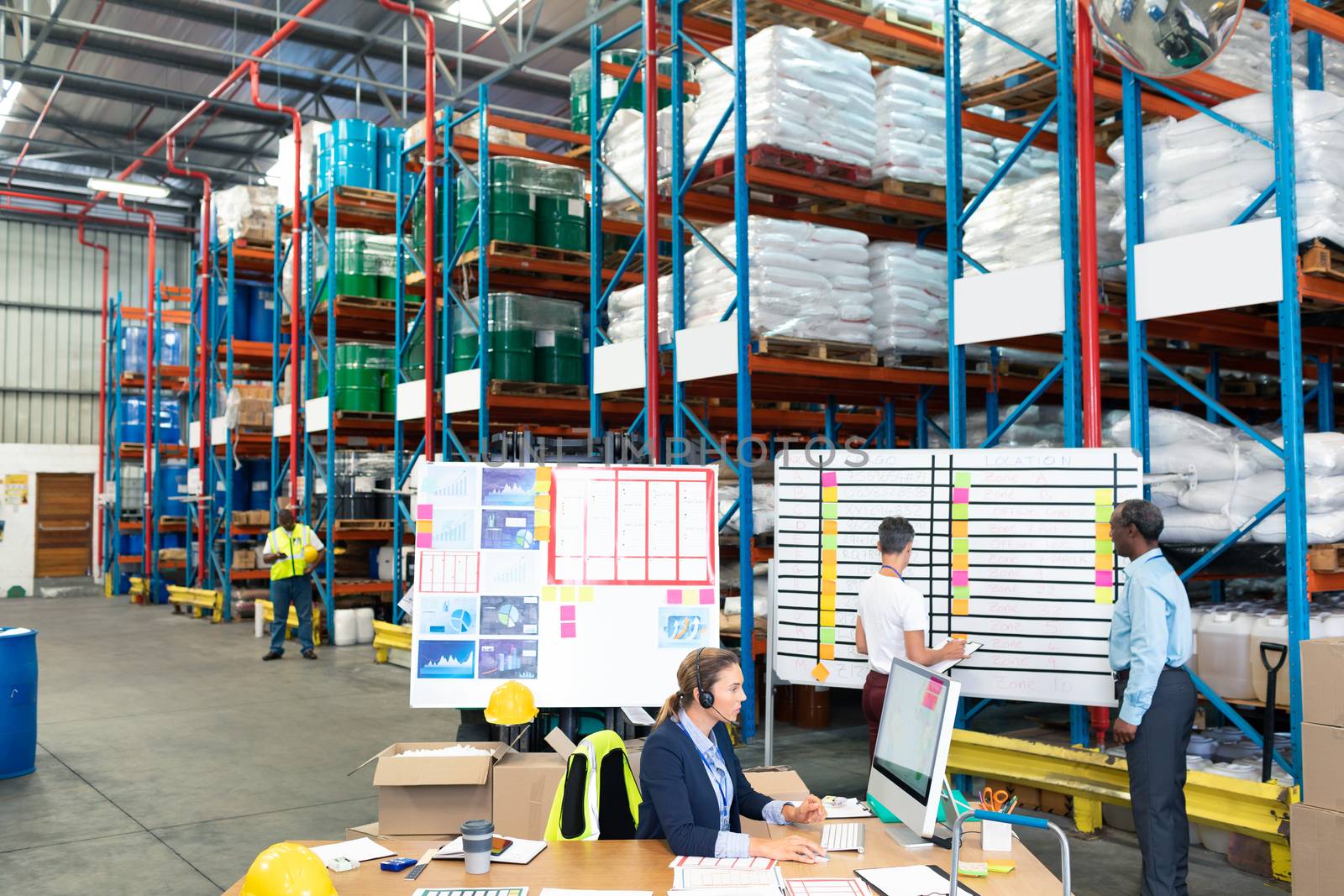 High angle view of Caucasian female manager working on computer while coworkers discussing over whiteboard in warehouse. This is a freight transportation and distribution warehouse. Industrial and industrial workers concept