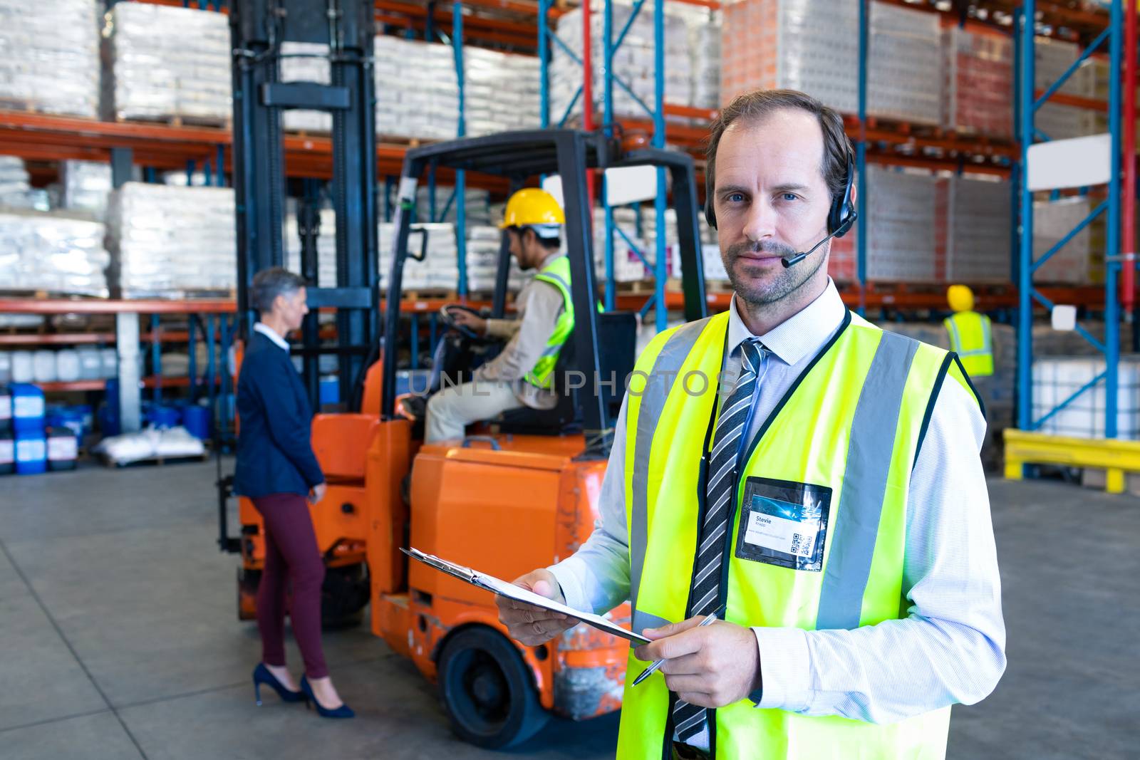 Front view of handsome Caucasian male supervisor with headset and clipboard looking at camera in warehouse. Diverse colleagues talking in the background. This is a freight transportation and distribution warehouse. Industrial and industrial workers concept
