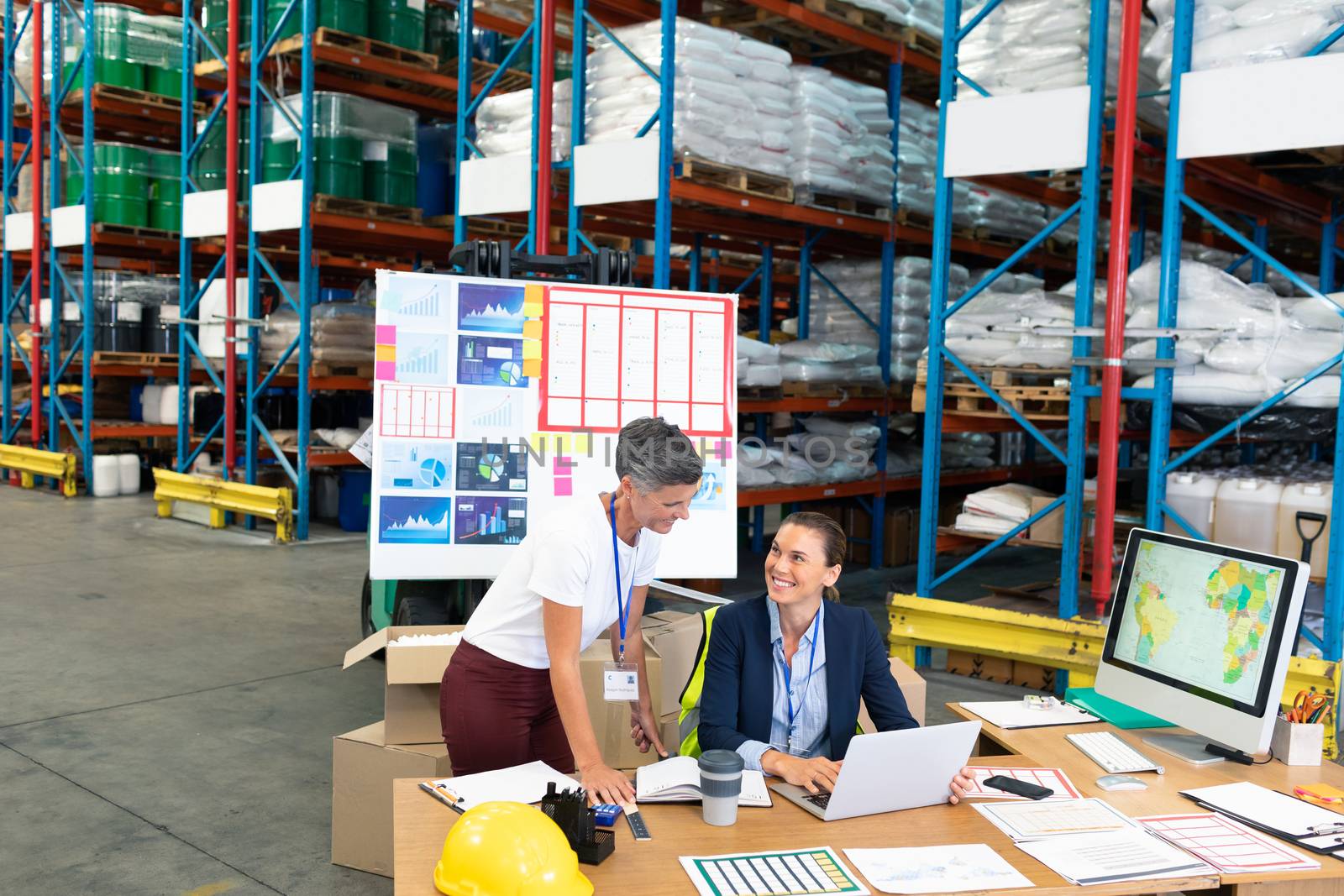 Front view of beautiful Caucasian female manager with her coworker discussing over laptop at desk in warehouse. This is a freight transportation and distribution warehouse. Industrial and industrial workers concept