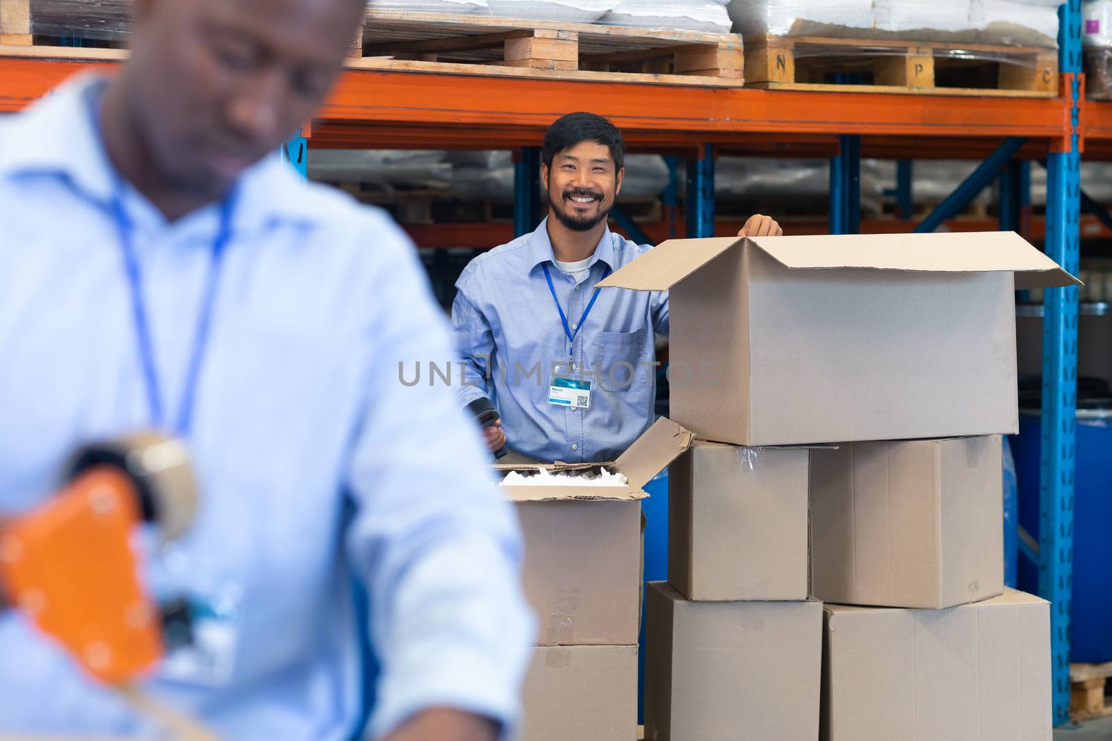 Front  view of happy mature Asian male staff looking at camera while checking stock in warehouse. On the foreground African-American male worker works in warehouse. This is a freight transportation and distribution warehouse. Industrial and industrial workers concept