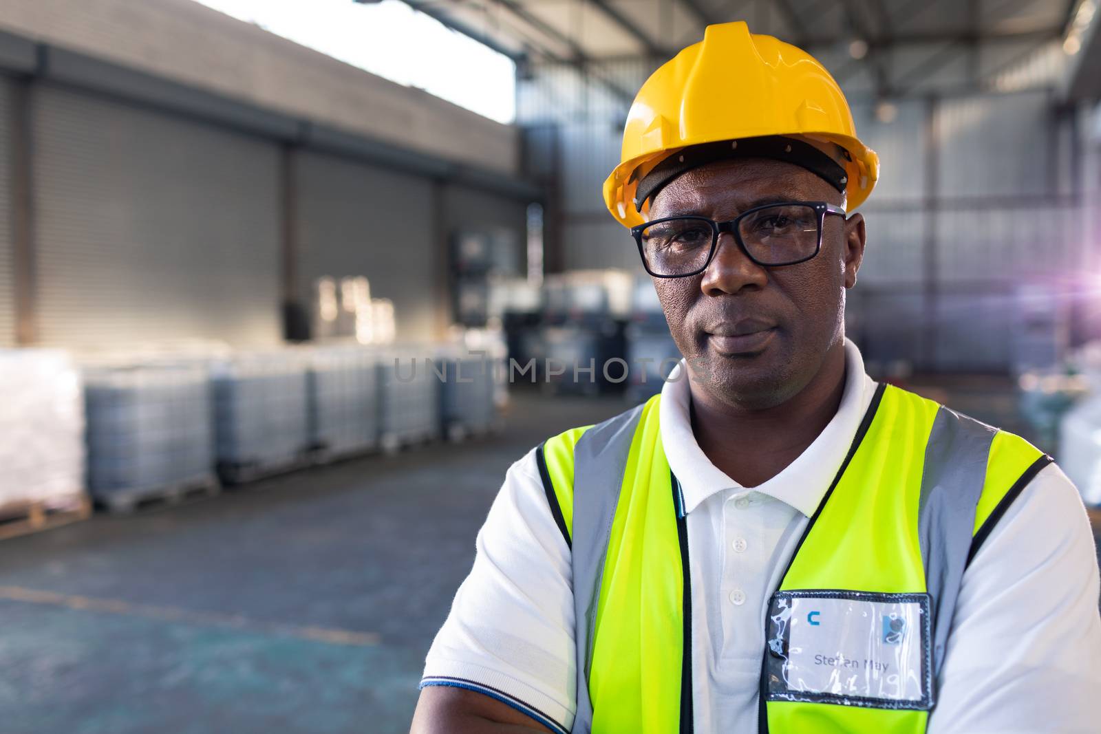 Portrait of African-american male staff in hardhat and reflective jacket standing with arms crossed in warehouse. This is a freight transportation and distribution warehouse. Industrial and industrial workers concept