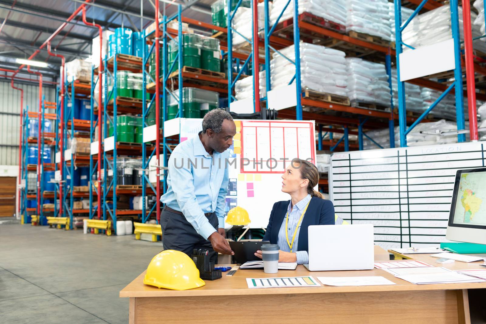 Front view of beautiful young Caucasian female manager with African-american coworker discussing over clipboard at desk in warehouse. This is a freight transportation and distribution warehouse. Industrial and industrial workers concept