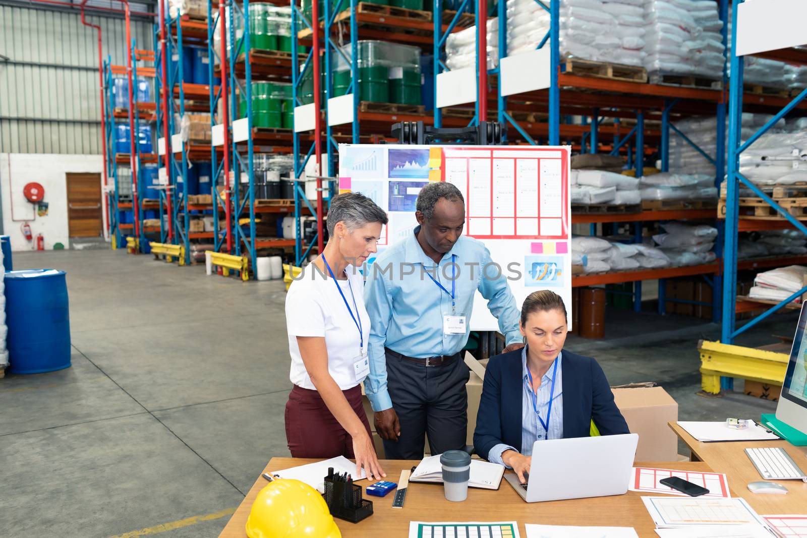 Front view of beautiful Caucasian female manager with her diverse coworkers discussing over laptop at desk in warehouse. This is a freight transportation and distribution warehouse. Industrial and industrial workers concept