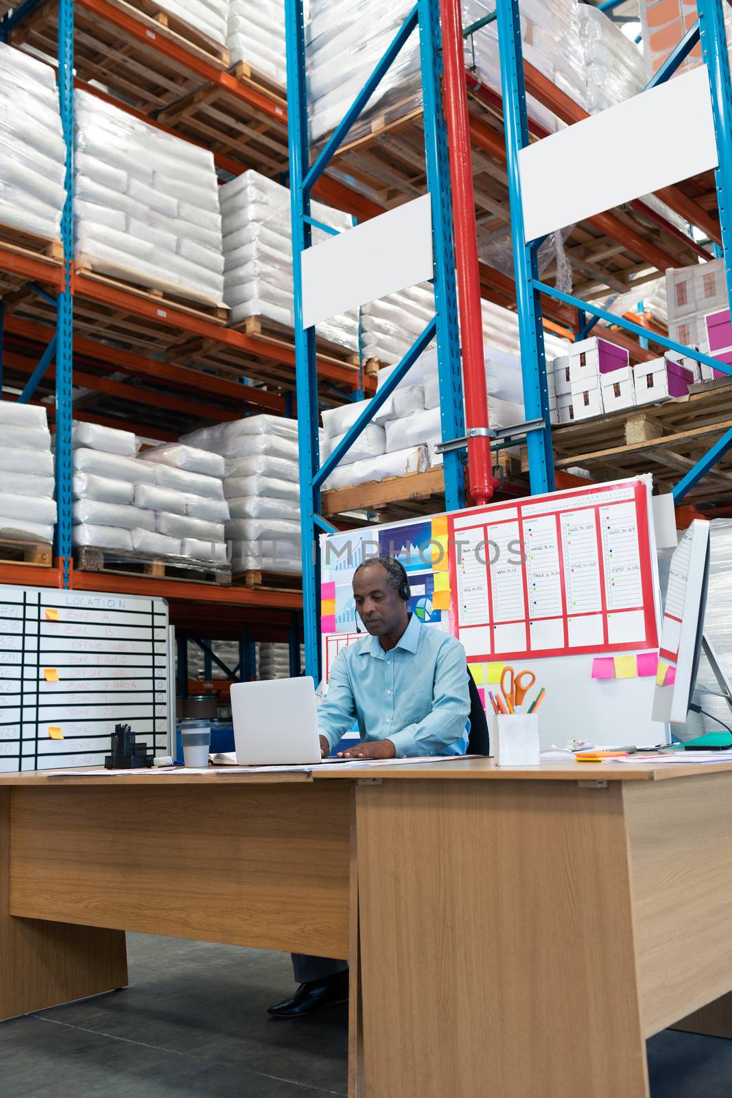Front view of handsome mature African-american male supervisor working on laptop at desk in warehouse. This is a freight transportation and distribution warehouse. Industrial and industrial workers concept