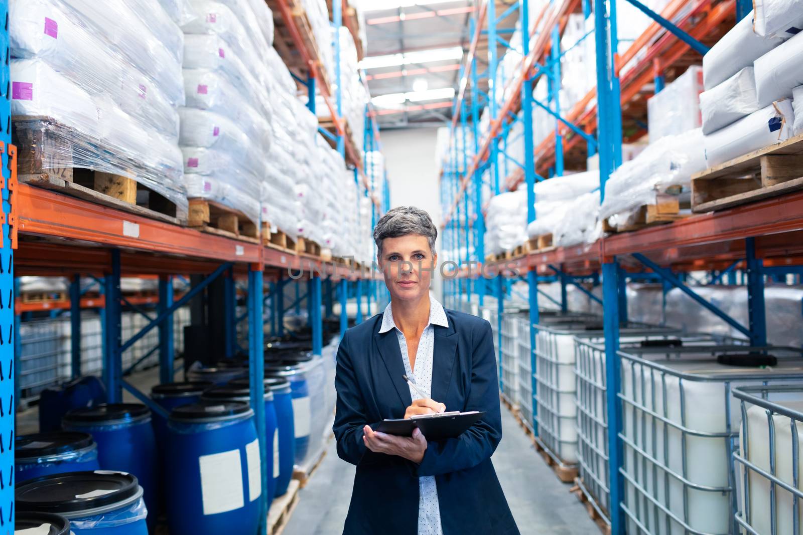 Portrait of beautiful mature Caucasian female manager looking at camera while writing on clipboard in warehouse. This is a freight transportation and distribution warehouse. Industrial and industrial workers concept
