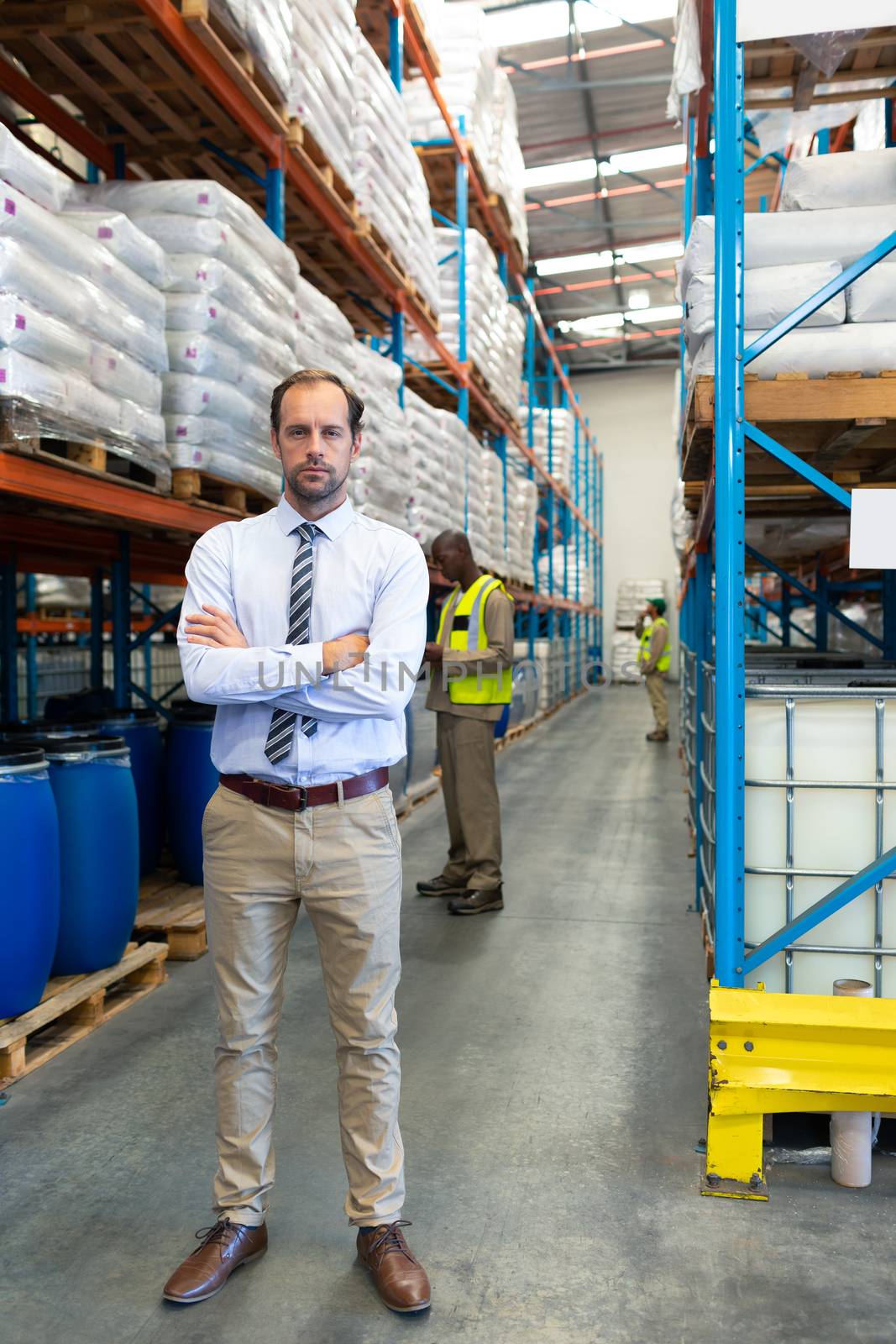 Front view of confident mature Caucasian male supervisor standing with arms crossed and looking at camera in warehouse. African-american colleagues working behind him. This is a freight transportation and distribution warehouse. Industrial and industrial workers concept