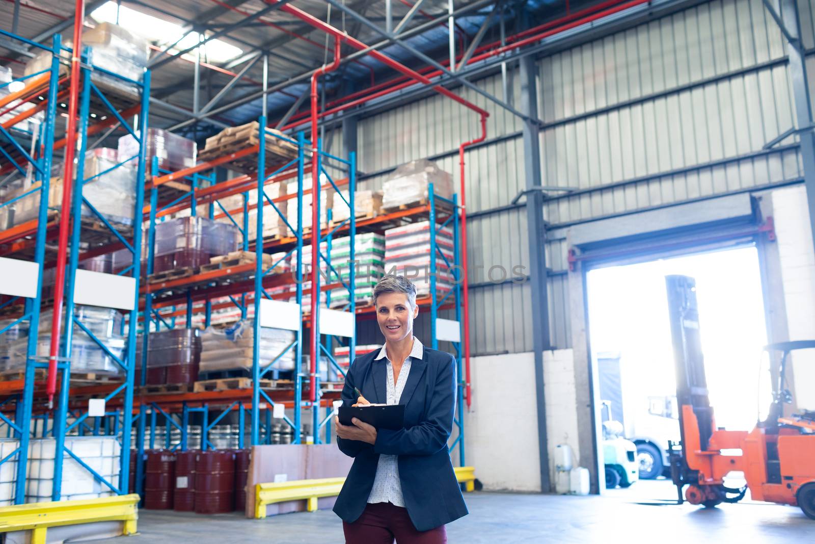 Front view of beautiful mature Caucasian female manager looking at camera while writing on clipboard in warehouse. This is a freight transportation and distribution warehouse. Industrial and industrial workers concept