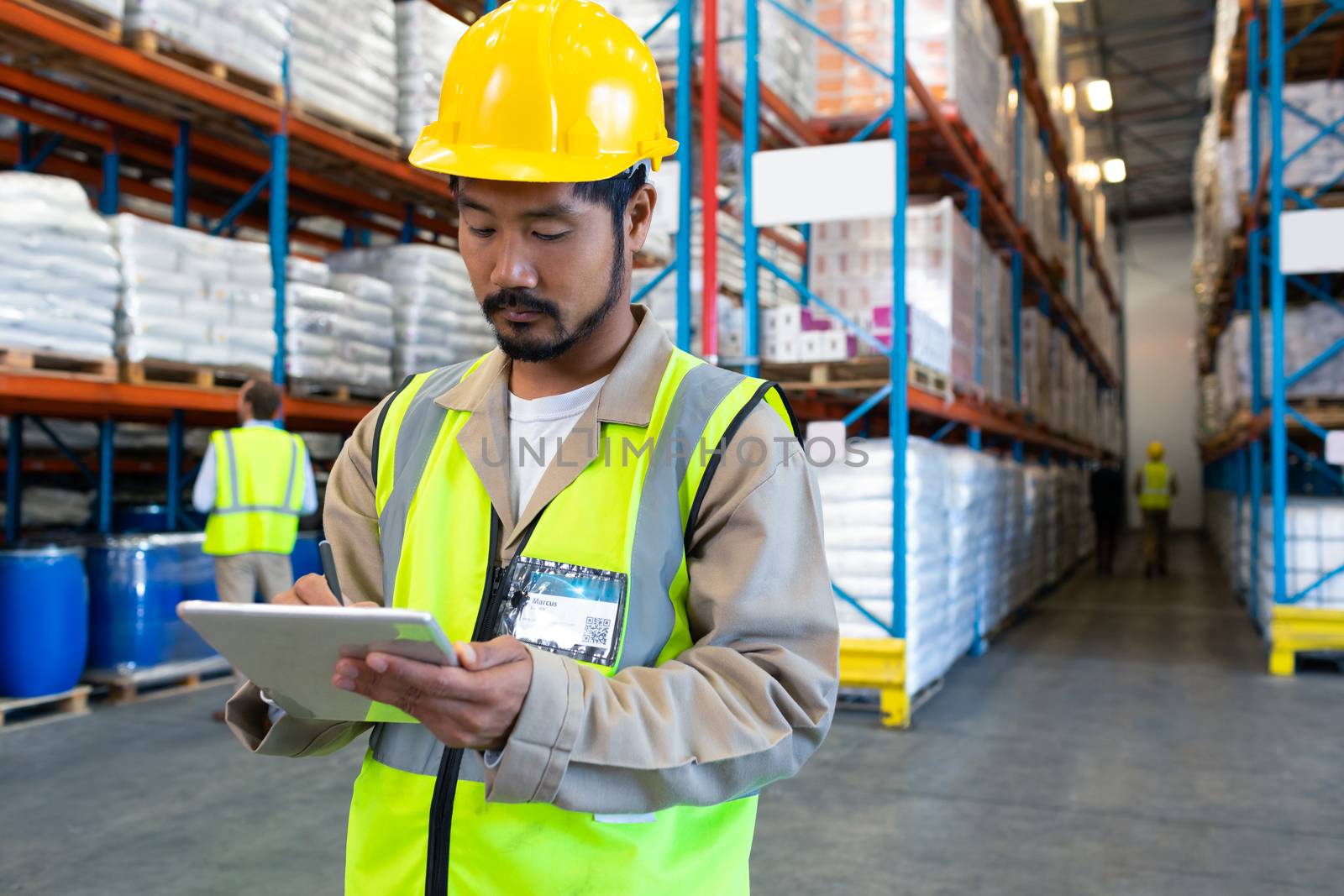 Front view of handsome mature Asian male worker working on digital tablet in warehouse. Diverse colleagues working in the background. This is a freight transportation and distribution warehouse. Industrial and industrial workers concept