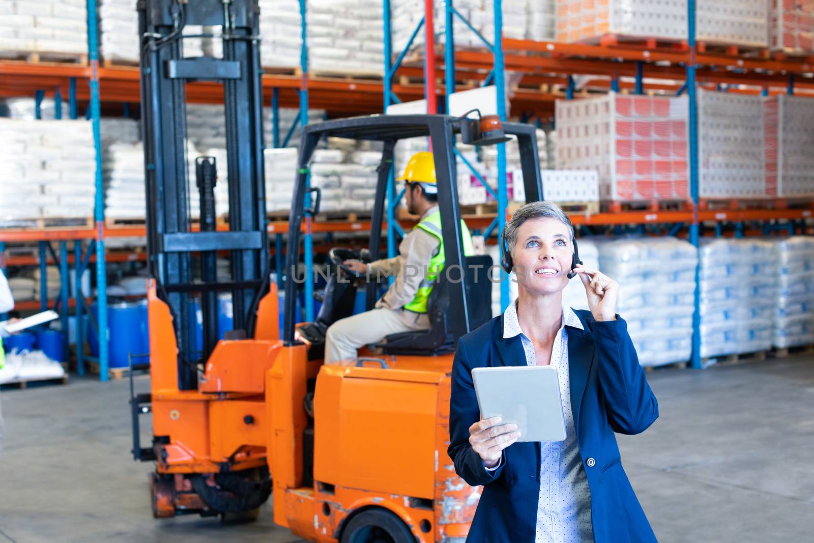 Front view of beautiful mature Caucasian female manager with digital tablet talking on headset in warehouse. Asian coworker sitting on forklift. This is a freight transportation and distribution warehouse. Industrial and industrial workers concept