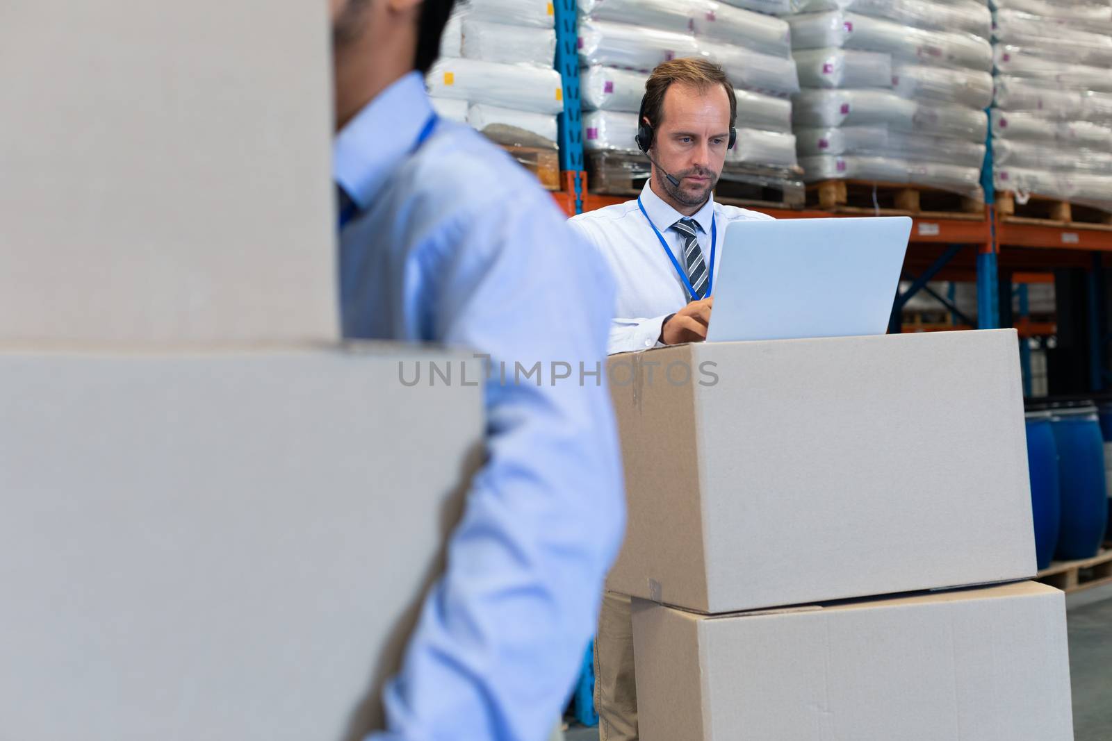 Front view of handsome mature Caucasian male supervisor with headset working on laptop in warehouse. Asian male worker holding cardboard boxes on the foreground. This is a freight transportation and distribution warehouse. Industrial and industrial workers concept