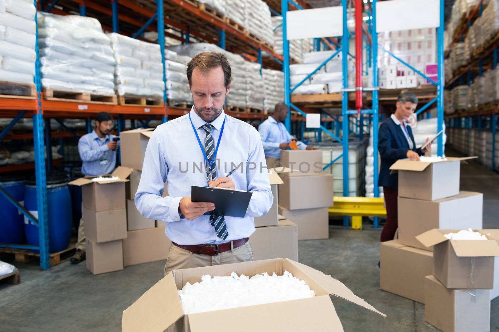 Front view of handsome mature Caucasian male supervisor writing on clipboard in warehouse. Diverse workers checking stock in cardboard boxes behind supervisor. This is a freight transportation and distribution warehouse. Industrial and industrial workers concept