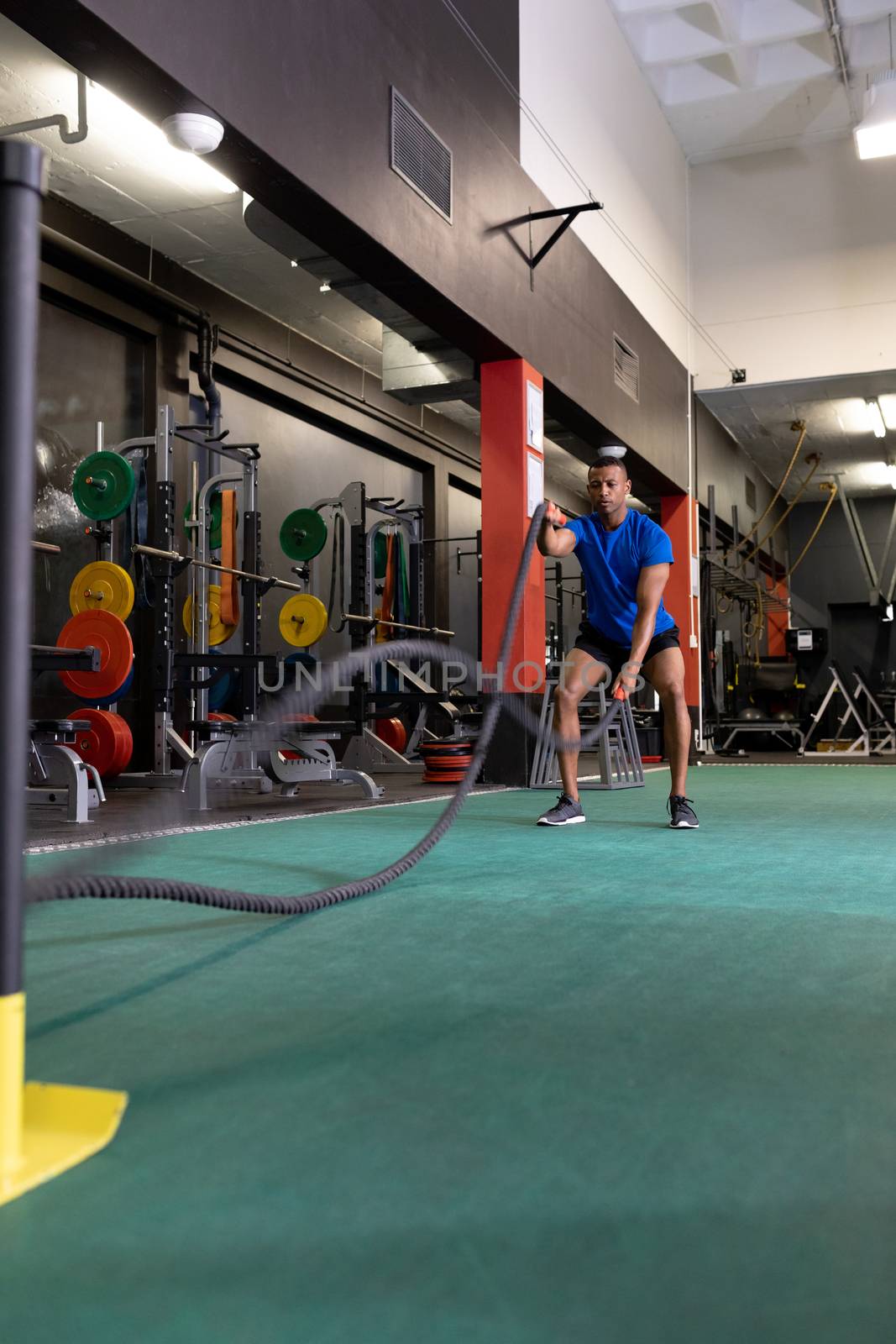 Front view of an African-American man doing battle rope exercises inside a room at a sports centre. Bright modern gym with fit healthy people working out and training. Bright modern gym with fit healthy people working out and training