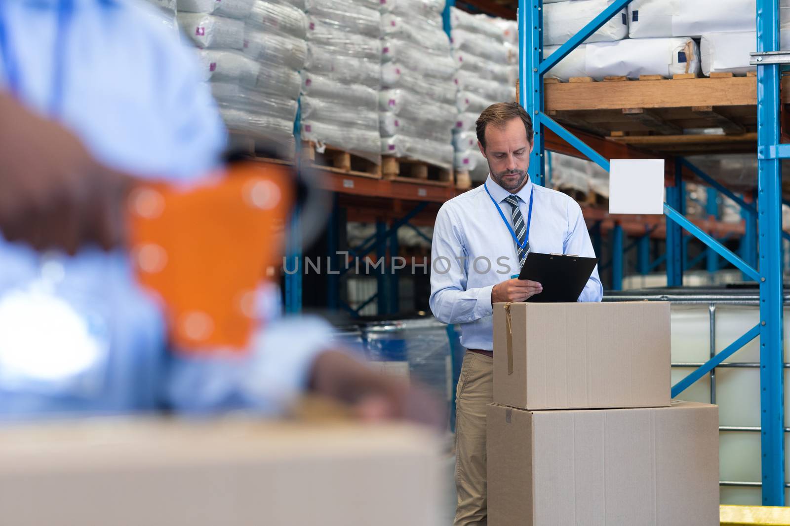 Front view of handsome mature Caucasian male supervisor working on clipboard while African-american staff works on the foreground in warehouse. This is a freight transportation and distribution warehouse. Industrial and industrial workers concept
