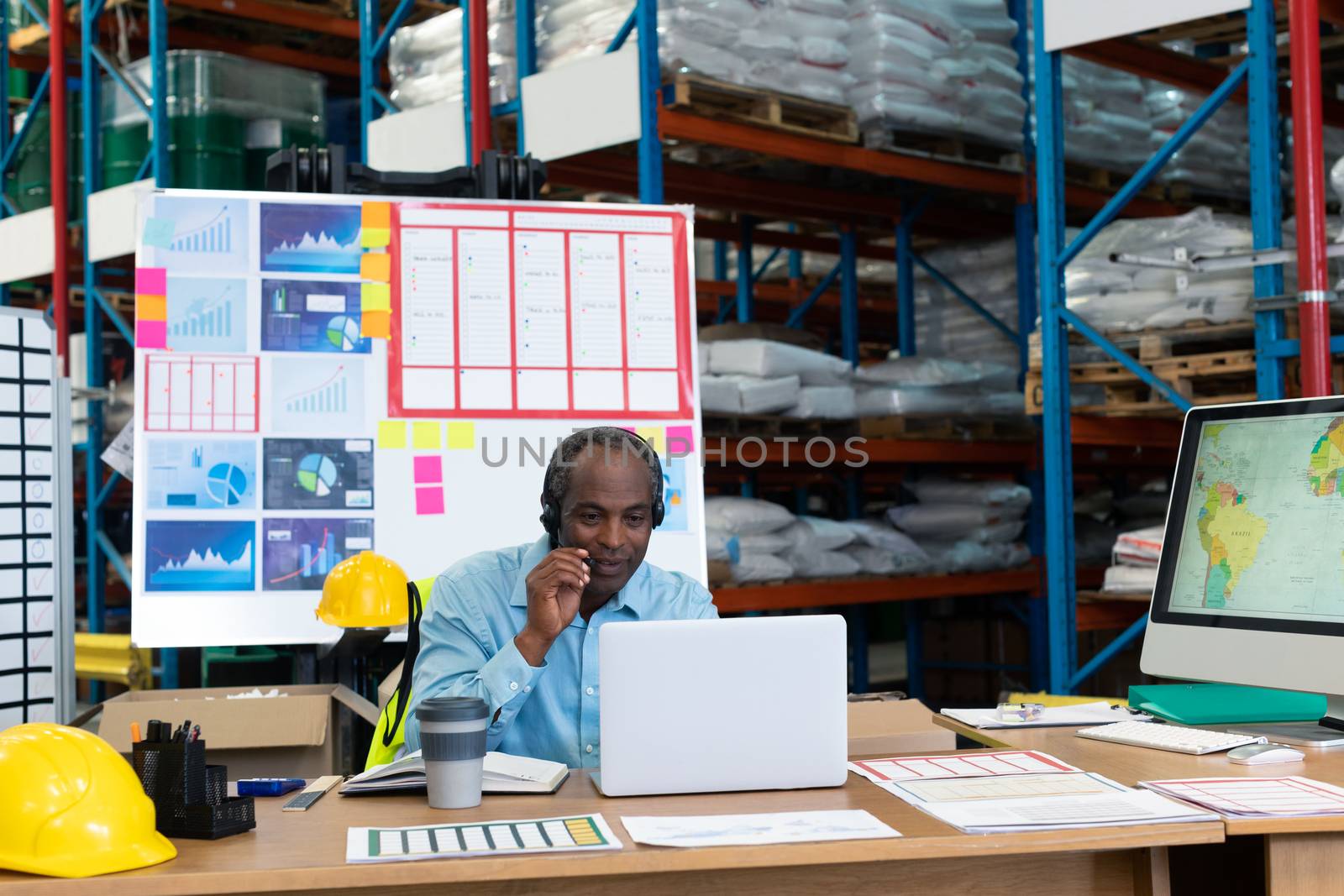 Front view of mature African american male supervisor talking on headset while using laptop at desk in warehouse. This is a freight transportation and distribution warehouse. Industrial and industrial workers concept