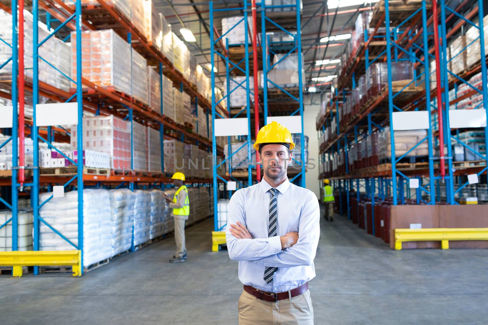 Front view of male supervisor standing with arms crossed and looking at camera in warehouse. This is a freight transportation and distribution warehouse. Industrial and industrial workers concept