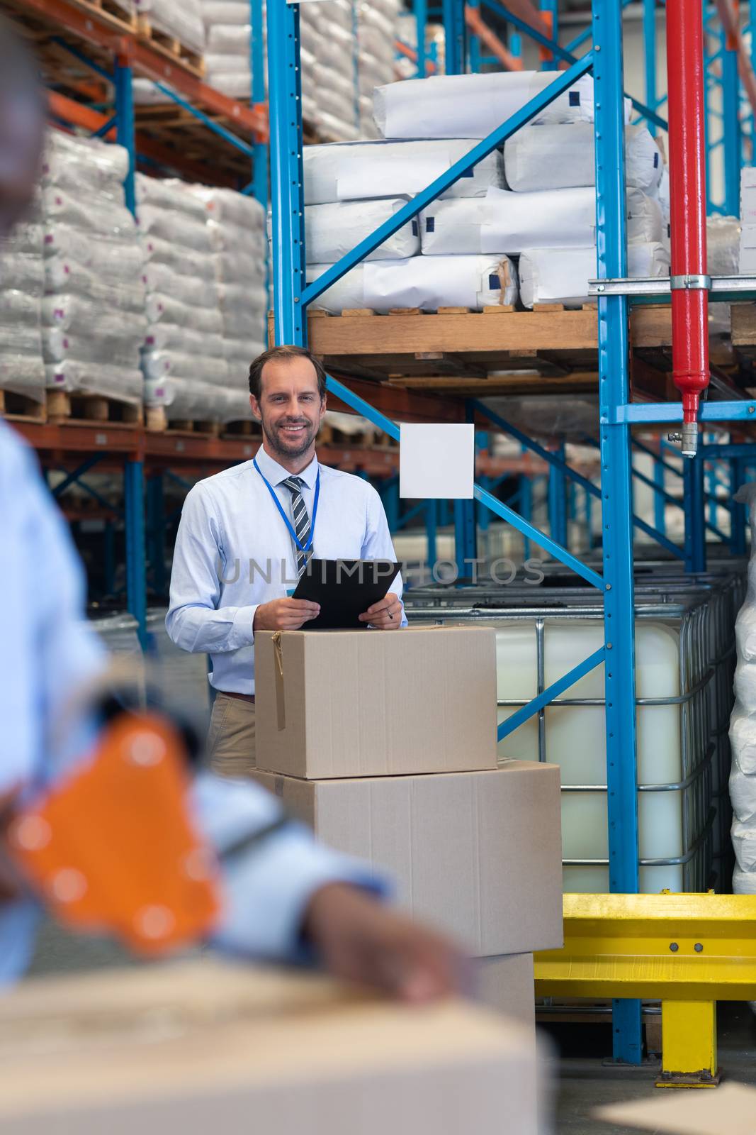 Front view of happy Caucasian male supervisor with clipboard looking at camera in warehouse. African-american staff works on the foreground. This is a freight transportation and distribution warehouse. Industrial and industrial workers concept