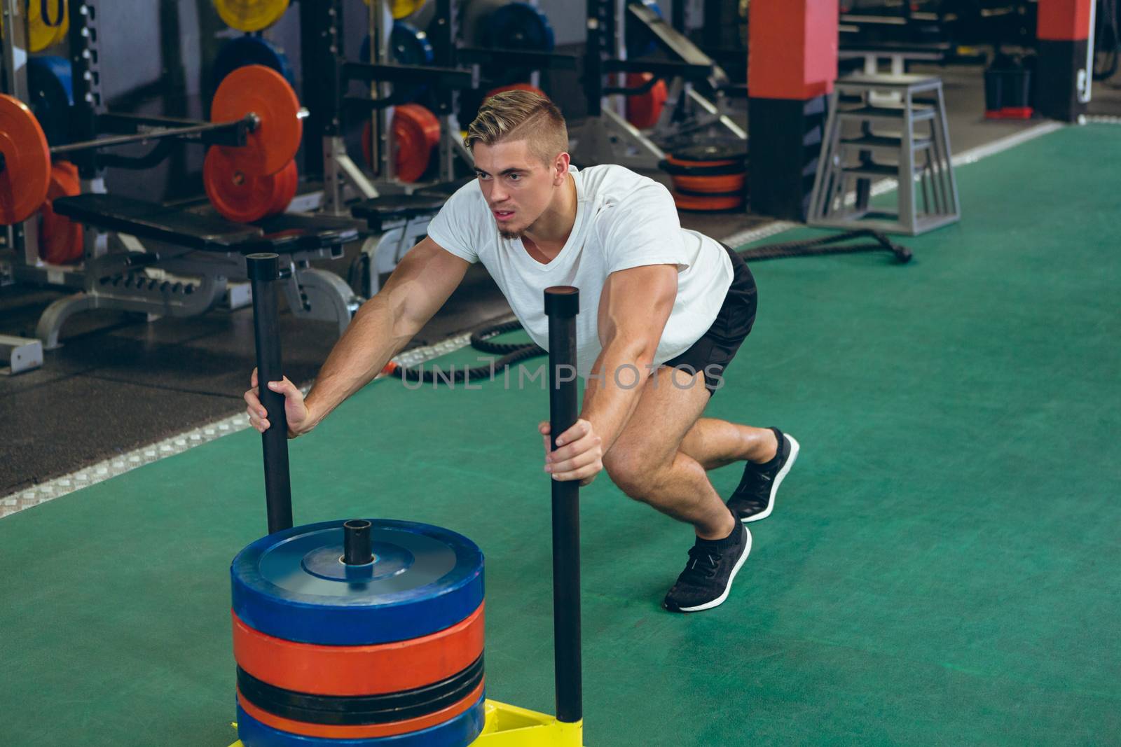 Front view of young Caucasian male athlete pushing prowler sled in fitness studio. Bright modern gym with fit healthy people working out and training