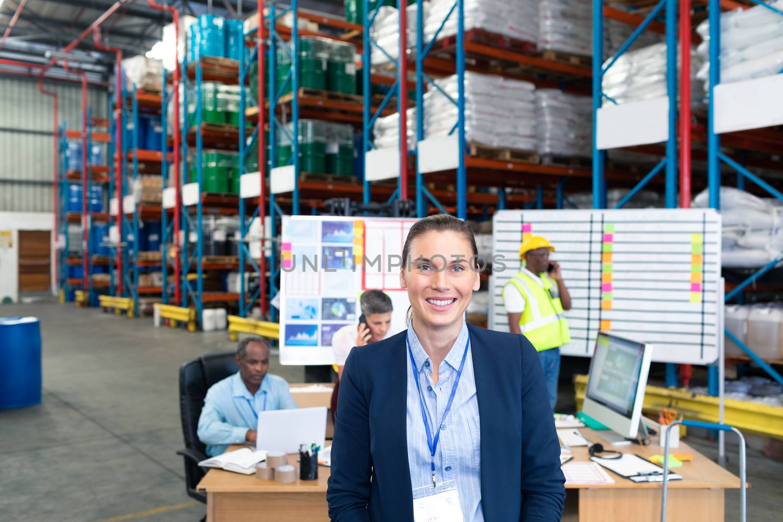 Portrait of happy mature Caucasian female manager looking at camera in warehouse. Diverse colleagues are working in the background. This is a freight transportation and distribution warehouse. Industrial and industrial workers concept