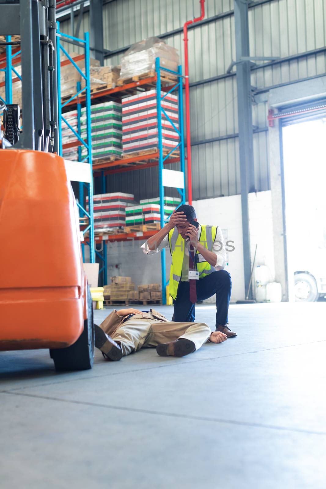 Front view of Caucasian Male supervisor talking on mobile phone while his coworker lying unconscious on the floor in warehouse. This is a freight transportation and distribution warehouse. Industrial and industrial workers concept