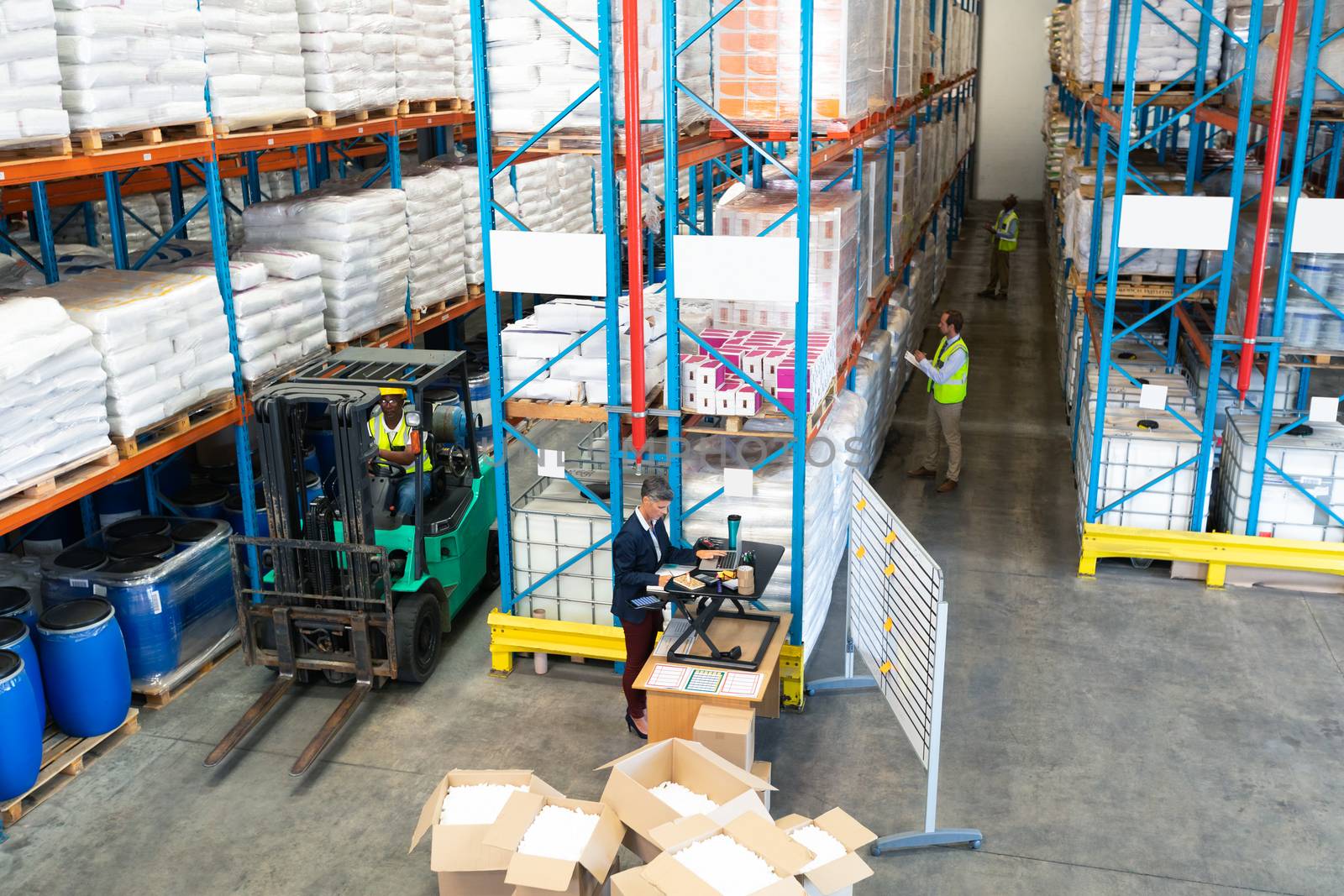 High angle view of mature diverse warehouse staff working together in warehouse. African-american worker driving a forklift in warehouse. This is a freight transportation and distribution warehouse. Industrial and industrial workers concept