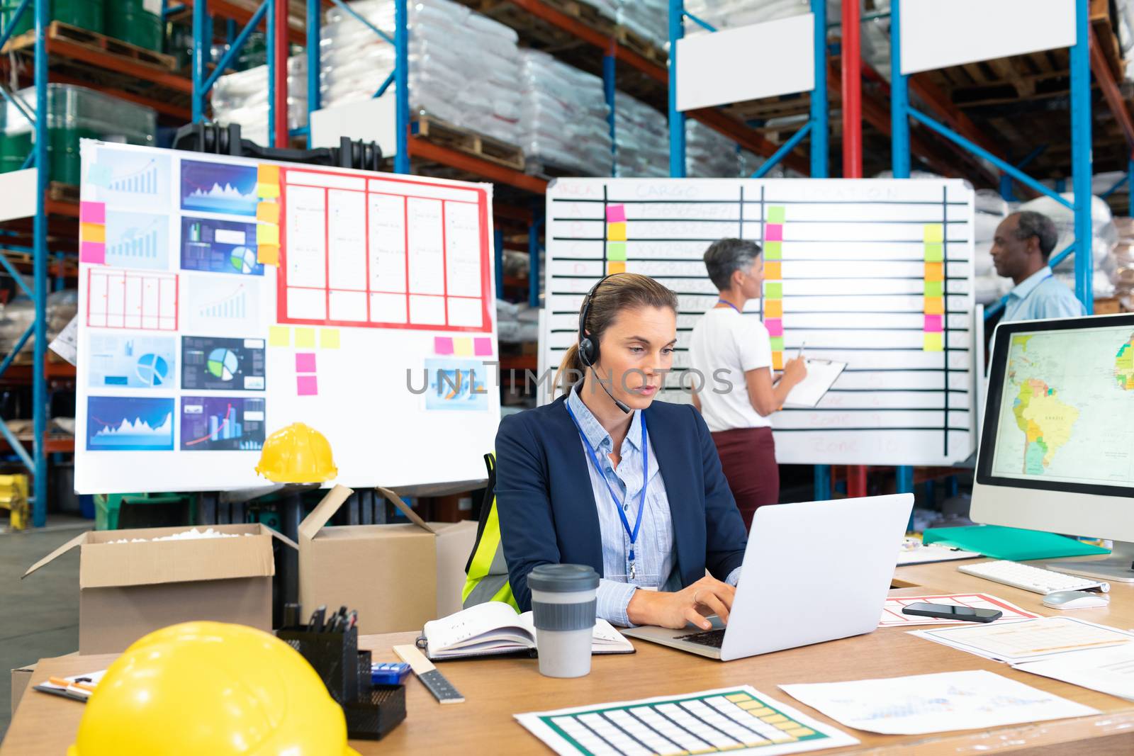 Front view of mature Caucasian female manager with headset using laptop at desk in warehouse. In the background diverse coworkers are discussing in front of whiteboard. This is a freight transportation and distribution warehouse. Industrial and industrial workers concept