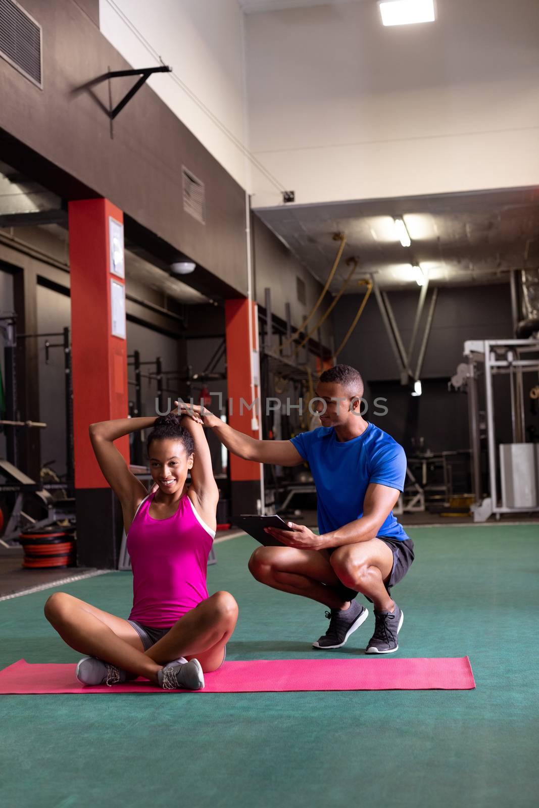 Front view of an African-American woman stretching while sitting crossed legged and an African-American man assists her inside a room at a sports center. Bright modern gym with fit healthy people working out and training. Bright modern gym with fit healthy people working out and training
