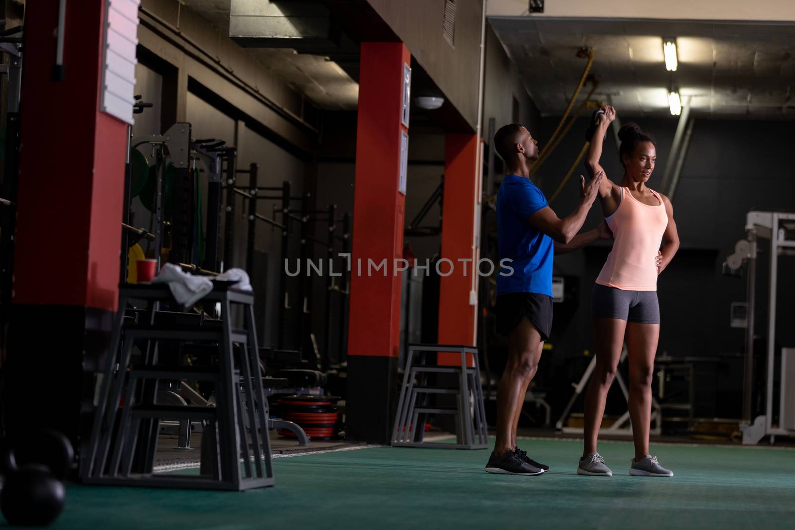 Front view of an African-American woman lifting ball weights and an African-American man assisting her inside a room at a sports centre. Bright modern gym with fit healthy people working out and training. Bright modern gym with fit healthy people working out and training