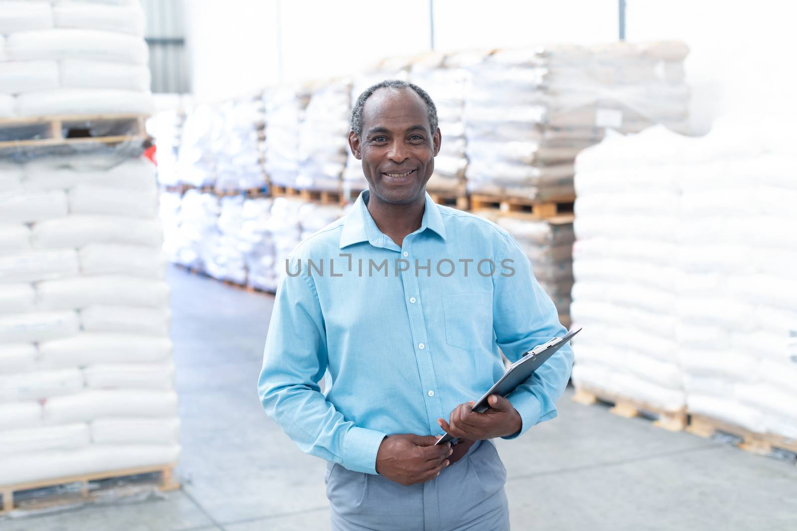 Portrait of happy mature African-american male supervisor with clipboard looking at camera in warehouse. This is a freight transportation and distribution warehouse. Industrial and industrial workers concept