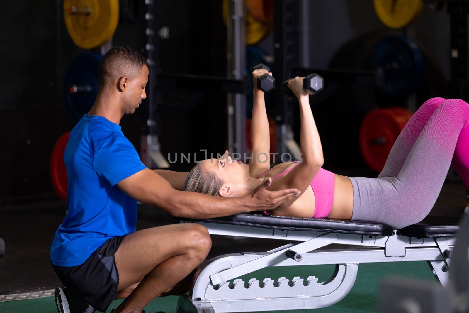 Side view of a Caucasian woman lifting dumbbells while lying on a gym bench and an African-American man assisting her inside a room at a sports centre. Bright modern gym with fit healthy people working out and training. Bright modern gym with fit healthy people working out and training
