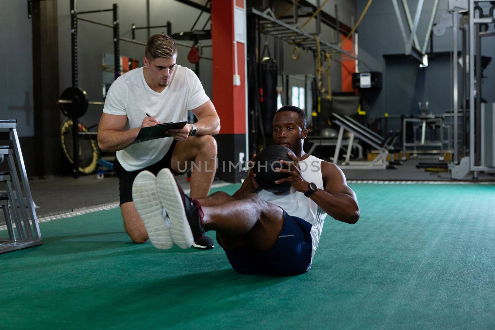 Front view of handsome young Caucasian male trainer writing on clipboard while young fit African-american male athletic exercising with exercise ball in fitness center. Bright modern gym with fit healthy people working out and training