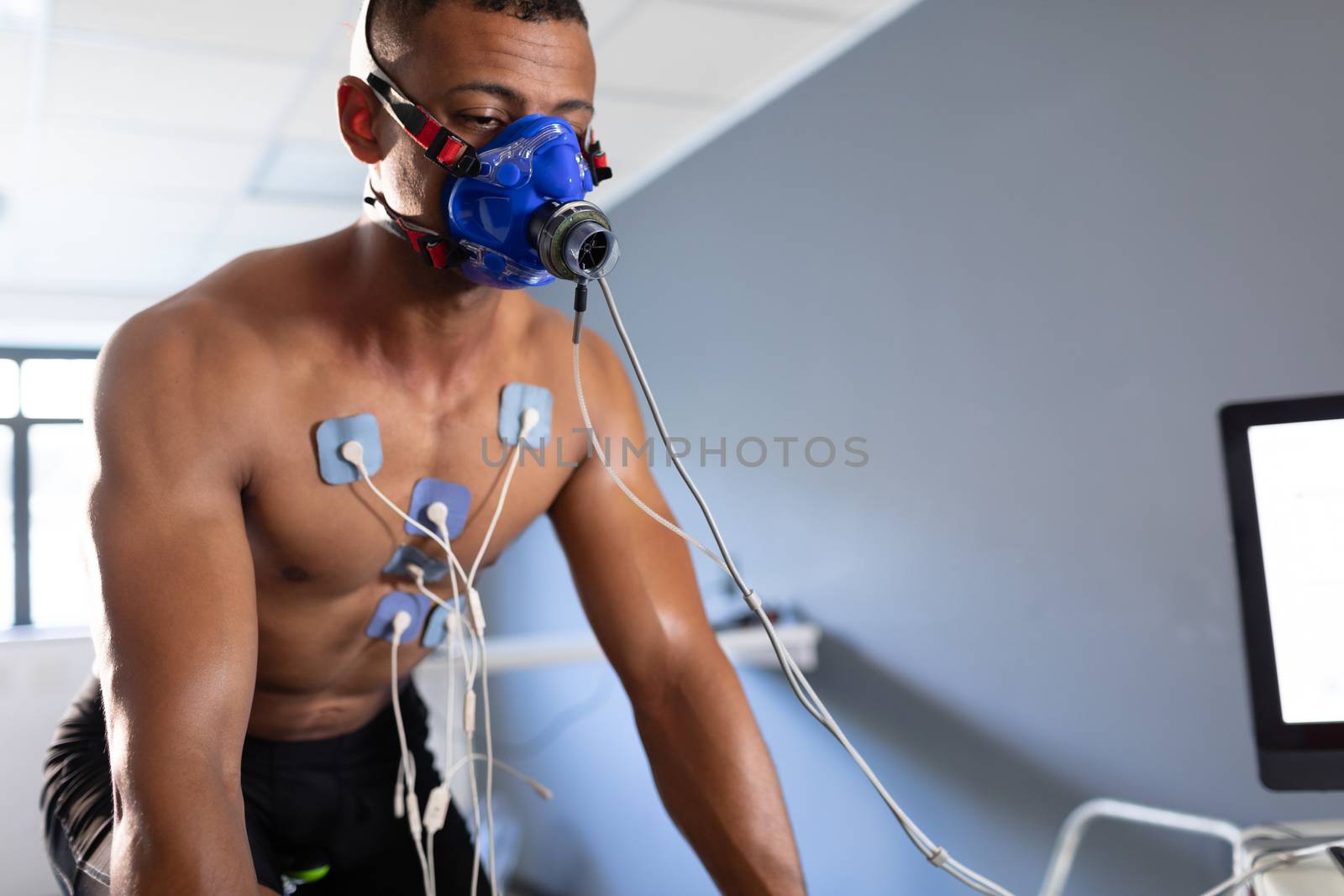Front view of a naked African-American athletic man doing a fitness test using a mask connected to a monitor while riding an exercise bike inside a room at a sports centre. Bright modern gym with fit healthy people working out and training. Athlete testing themselves with cardiovascular fitness test on exercise bike